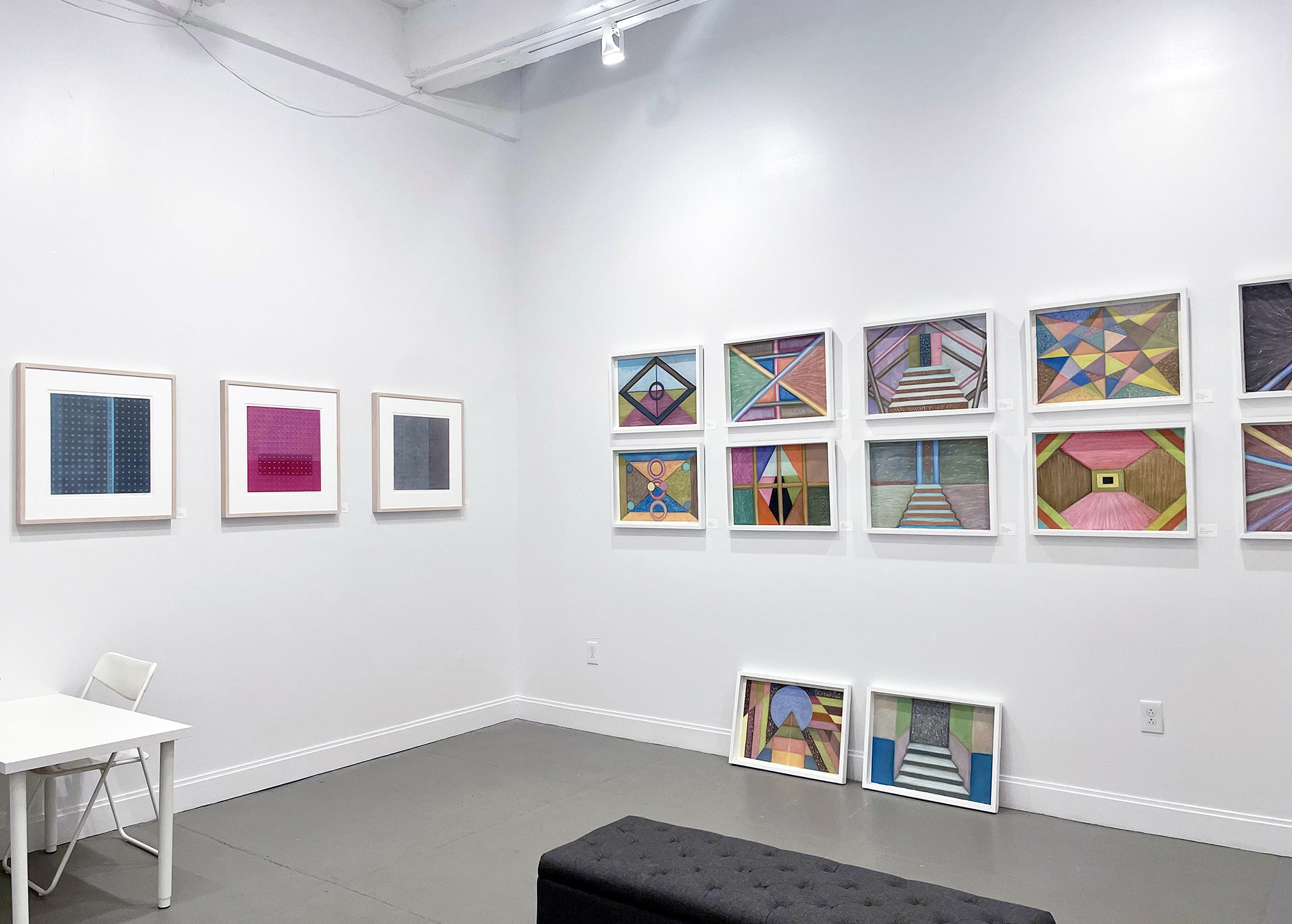 Bright, white-walled gallery space where on the left wall, three artworks hang side-by-side, and on the opposite right wall, two rows of three artworks each hang in two rows. Underneath, two more artworks rest on the floor.