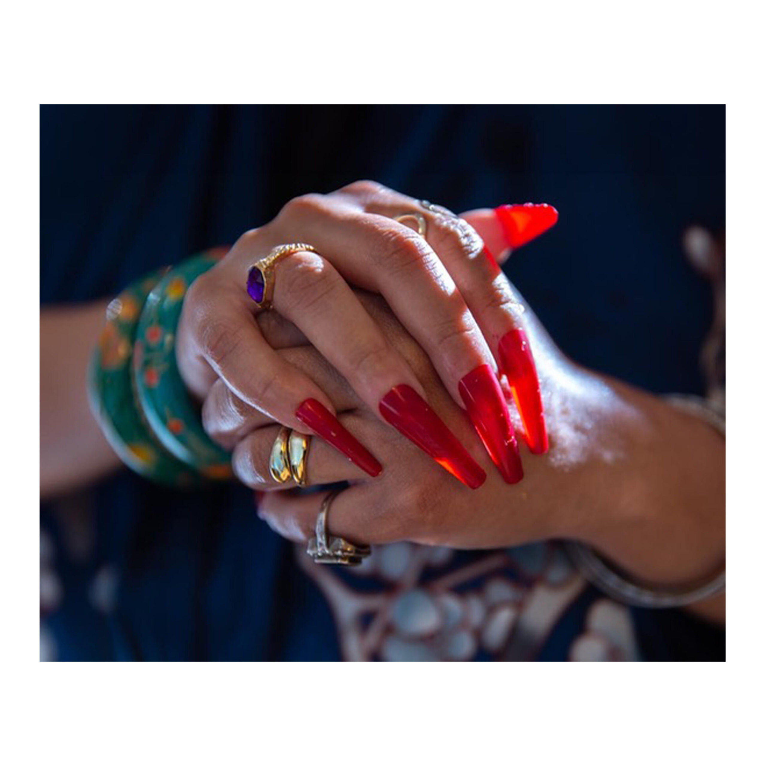 Close-up of clasped hands; the subject has long, bright red nails and their fingers are adorned with rings.