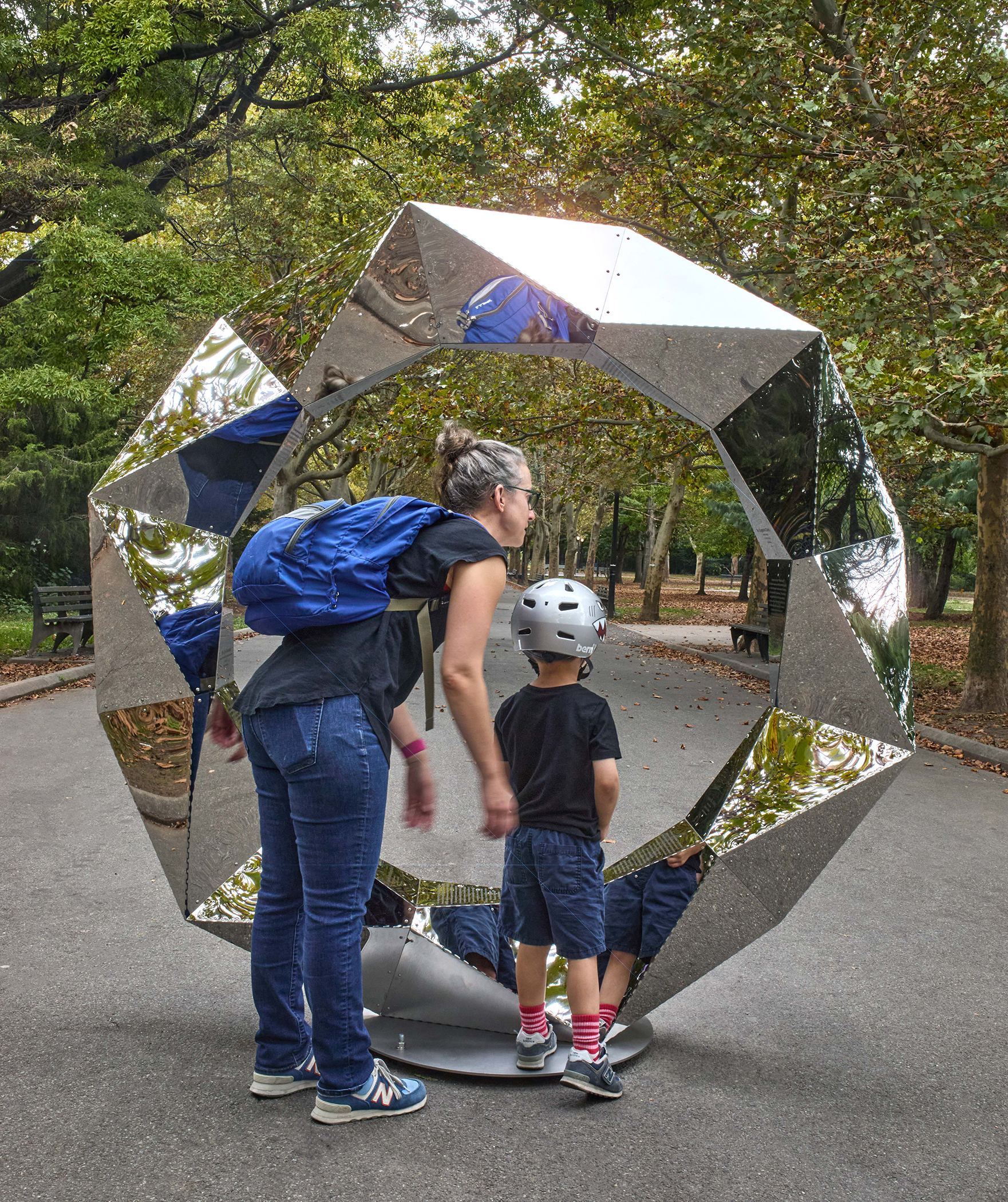 A woman and child in a park looking at a circular reflective sculpture that has geometric, paneled edges.