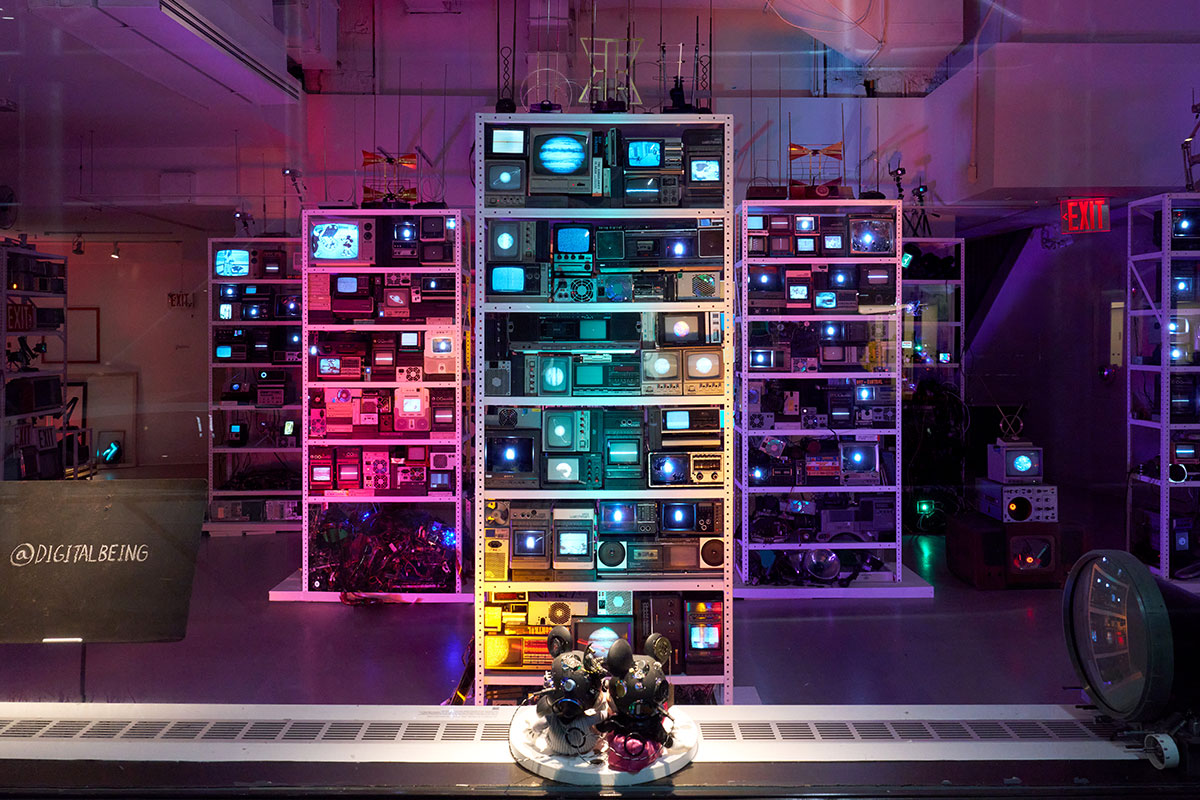 View through a glass window of a purple-lit room where several, tall bookshelf-like structures are packed with obsolete TVs and media players and lit with colored LEDs. On the left, a music stand with the text “@DIGITALBEING” is visible.