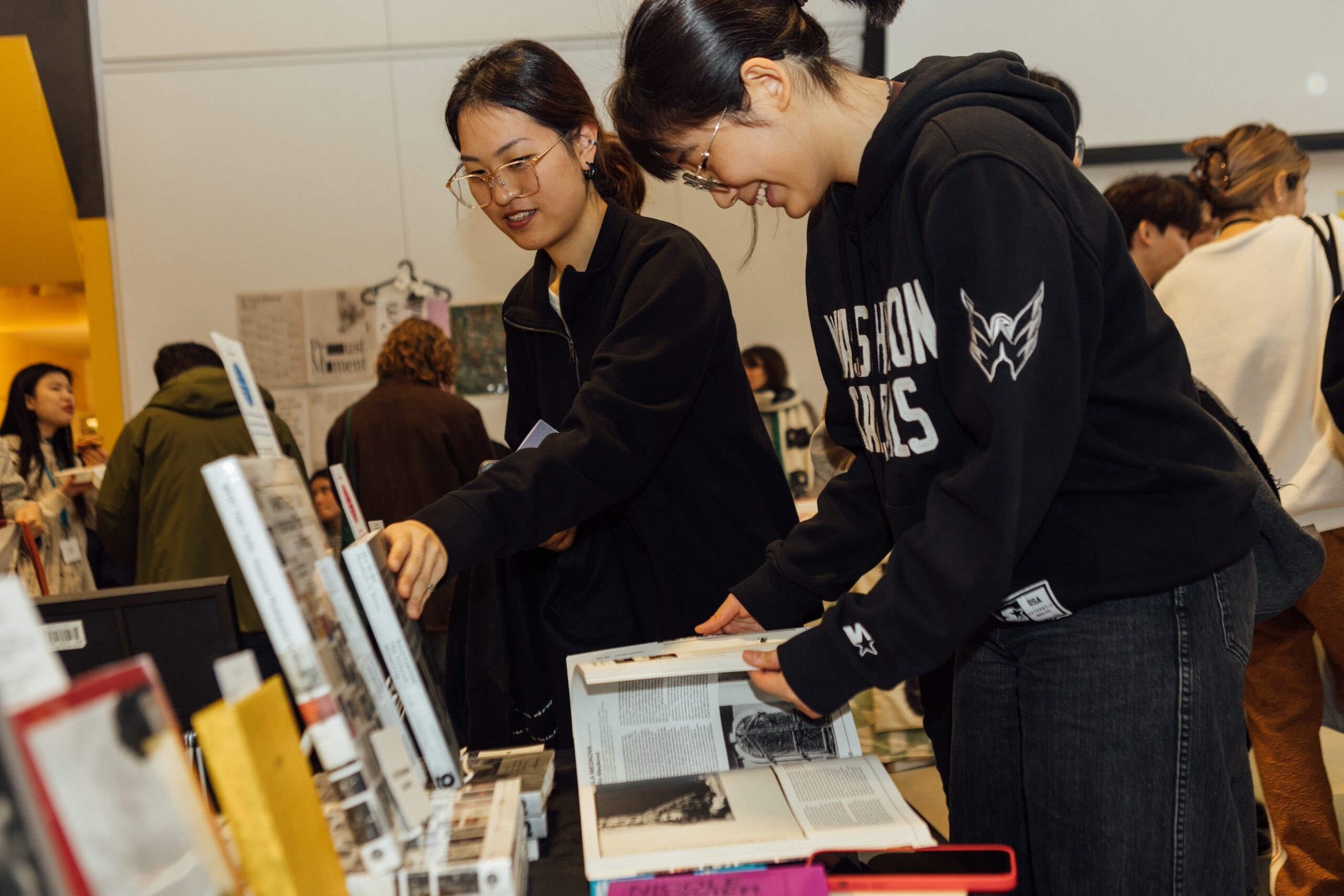 One attendee smiles, paging through a book at a table arranged with printed materials, while someone else reaches for another book on a display