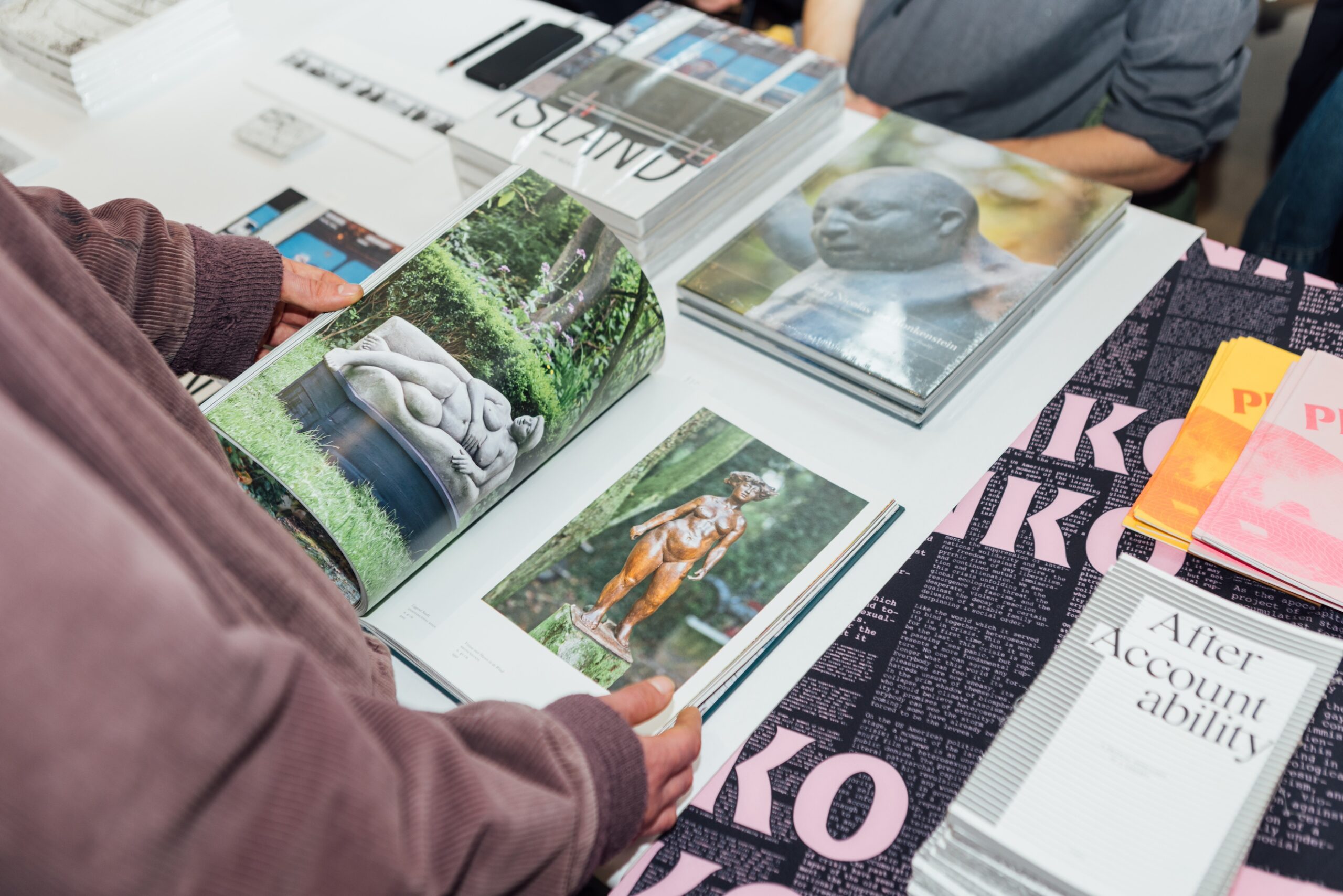 Someone pages through a magazine depicting sculptures of women at a table arranged with stacks of magazines and zines