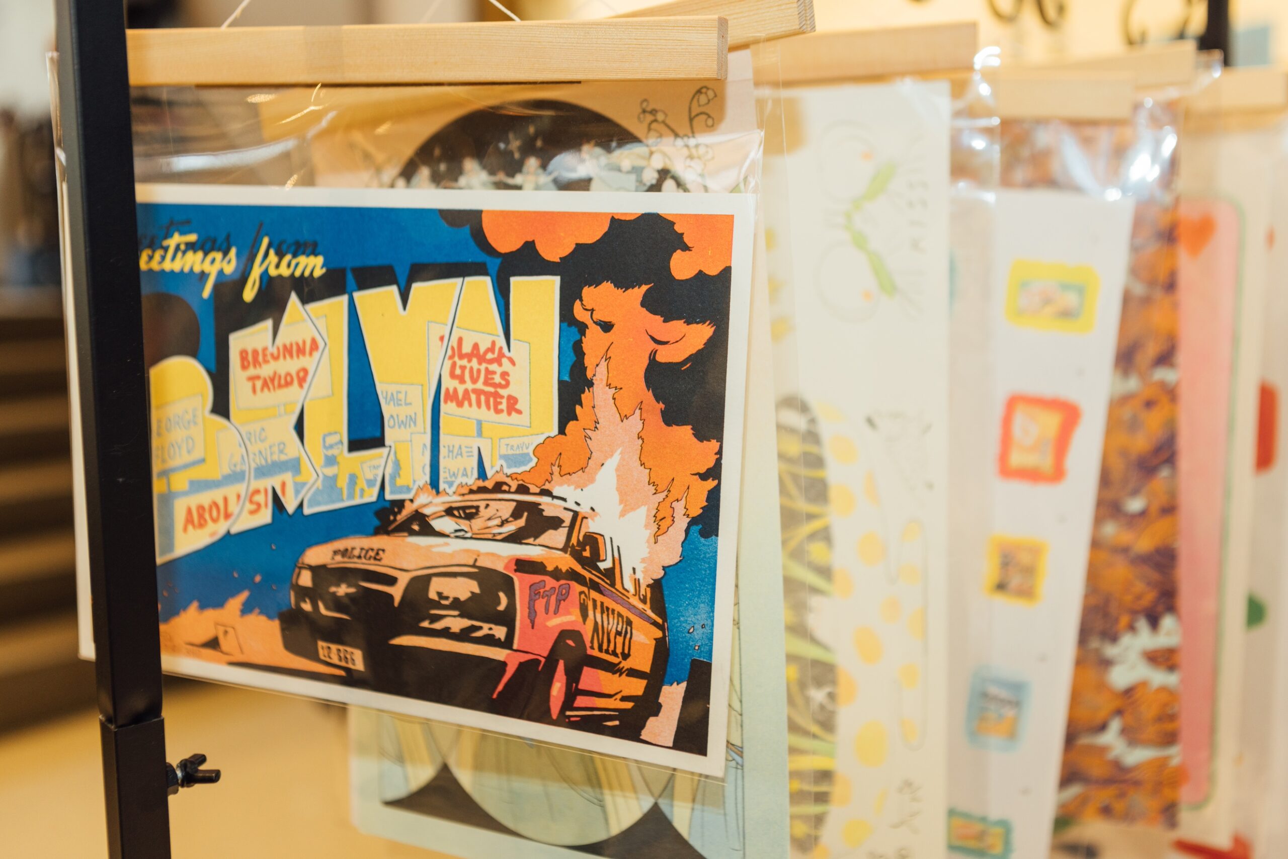 Detail of display depicting an illustrated “Greetings from BKLYN” postcard with imagery of a NYPD police car on fire and Black Lives Matter posters
