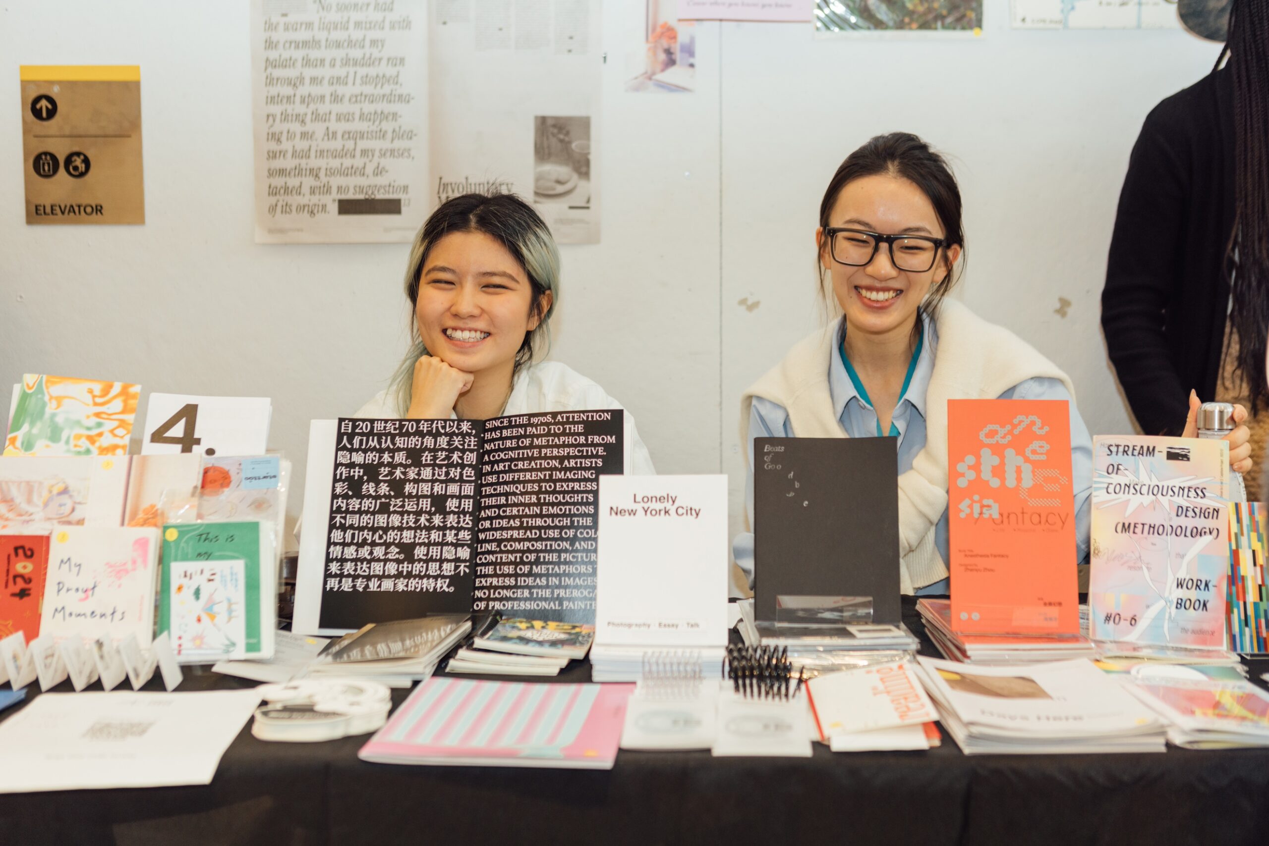 Two vendors smile behind a table displaying zines, looking at the camera