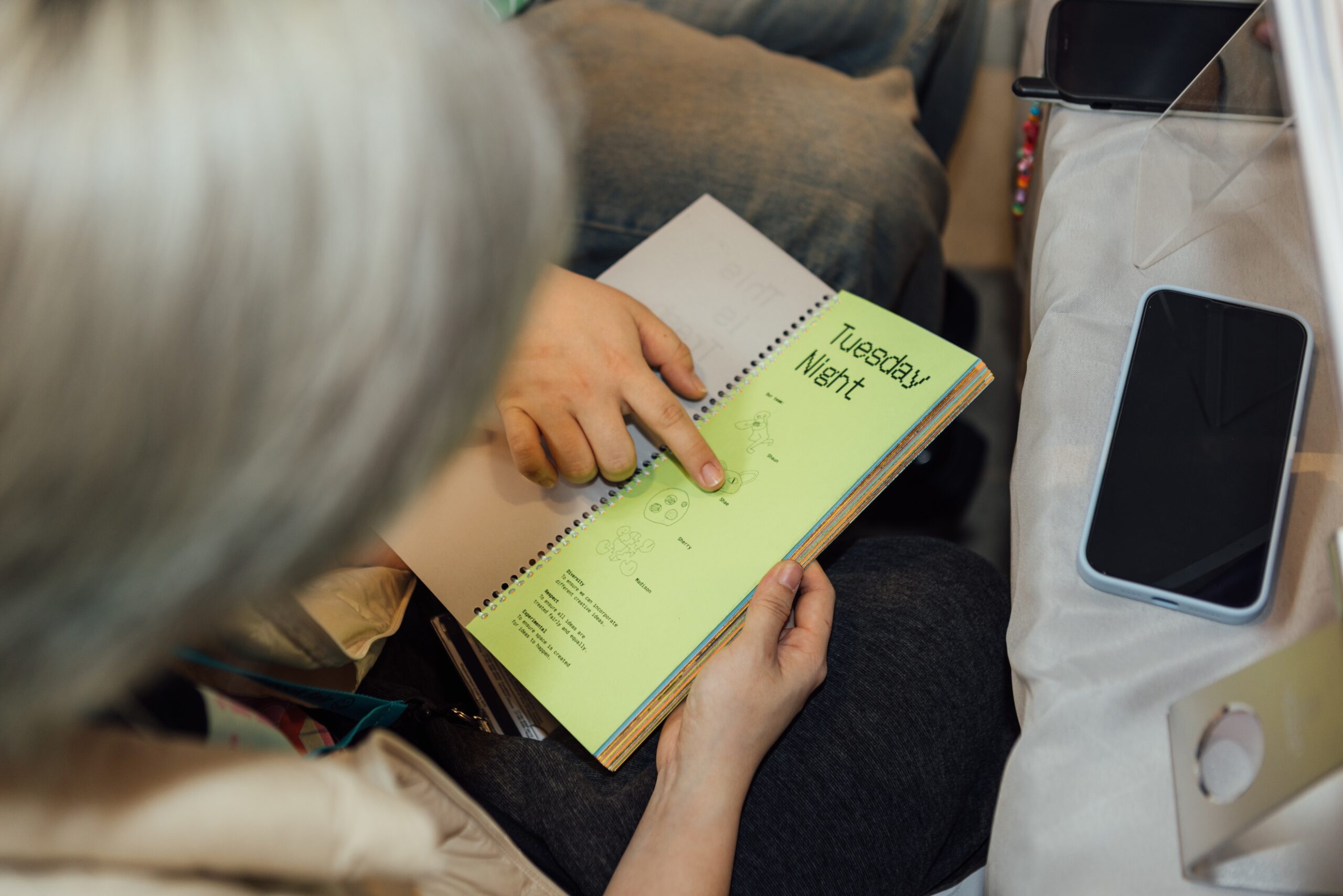 Close-up of two people looking at a spiral-bound booklet with pastel green paper titled “Tuesday Night”