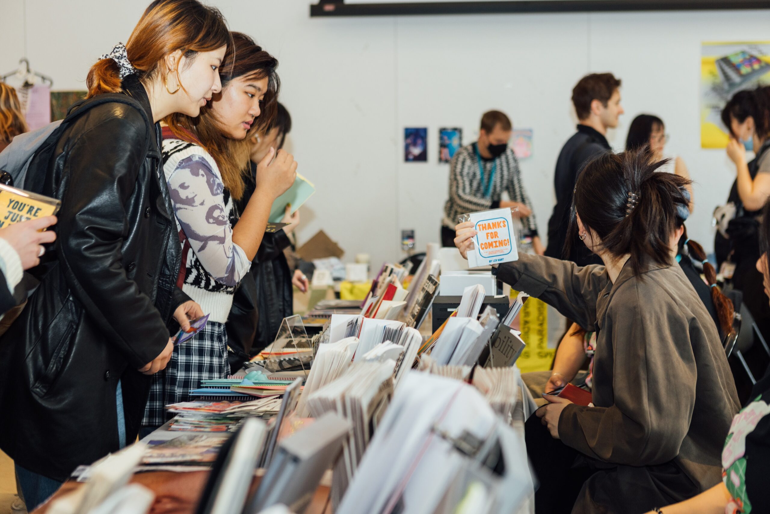 Fair attendees stand in front of tables arranged with printed materials, looking over at a vendor who holds up a card that says “Thanks for Coming”