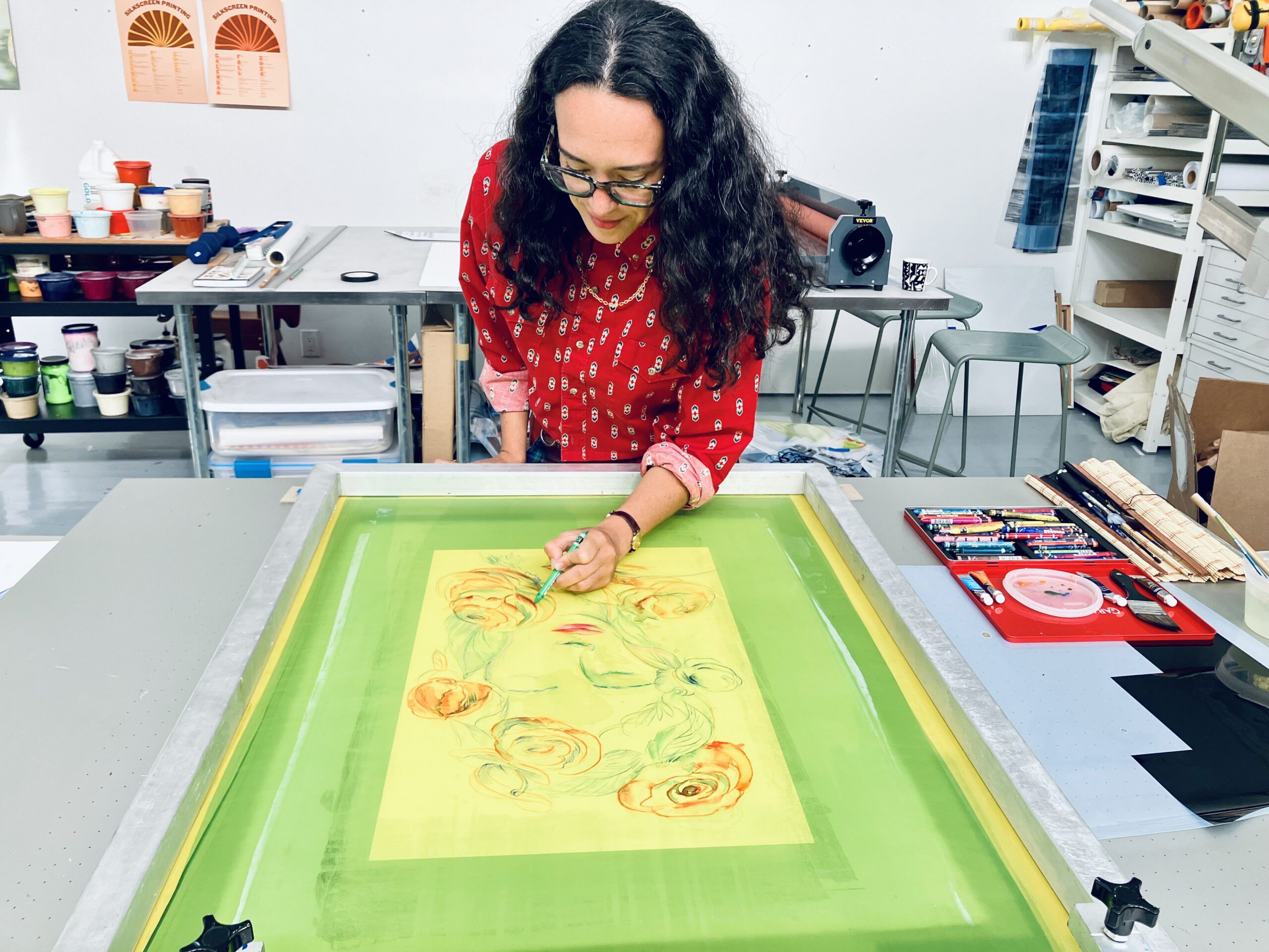 Artist with glasses and a bright red long-sleeved shirt sketches a design on a large, bright green printing screen. The room she is in is brightly lit with white walls; behind her is a cart with different colored inks next to a long silver table.