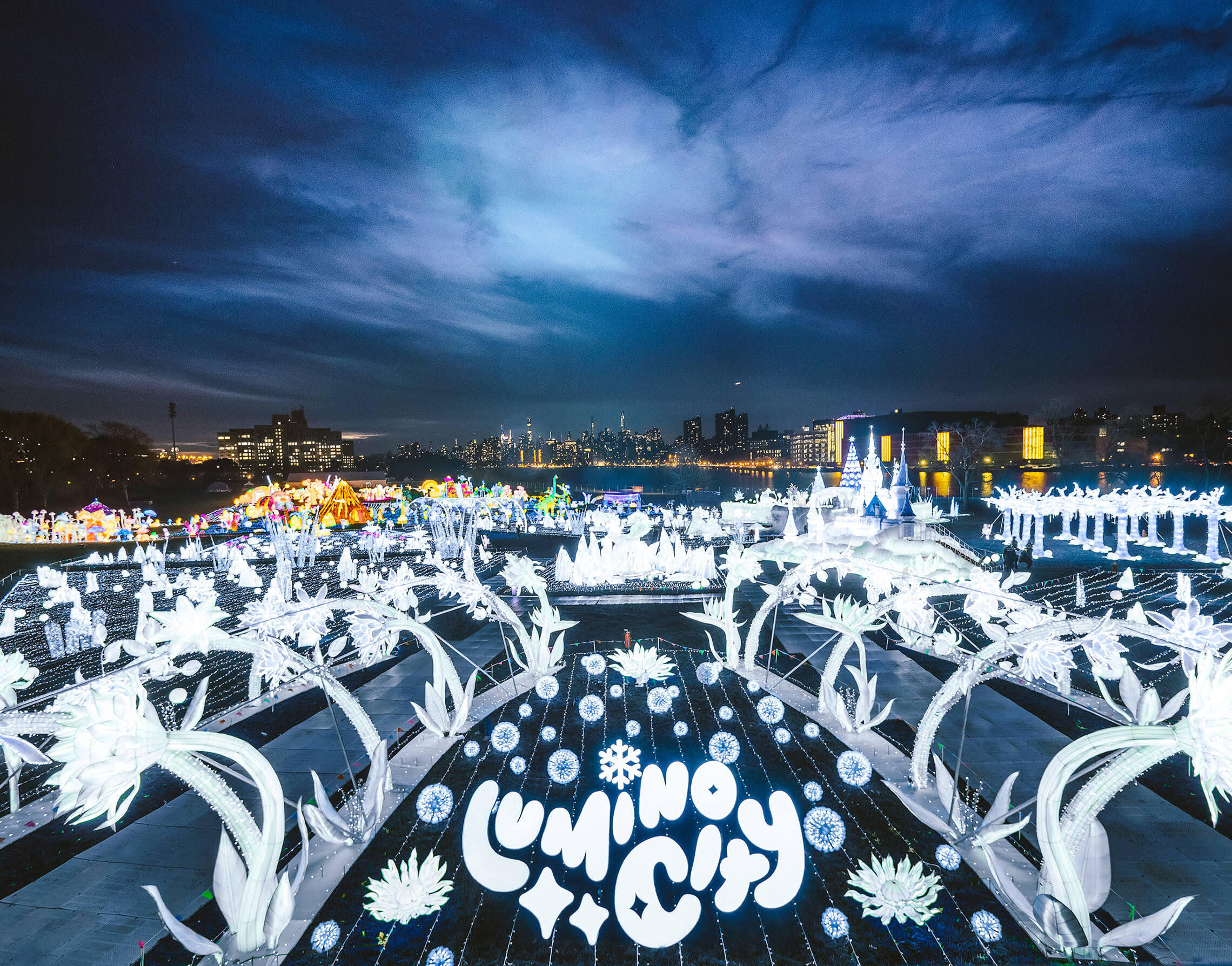 Festive winter outdoor light show, made up of bright white and colored light installations, at dusk with the New York skyline visible across the river. The sky is a dark blue. Bubble text at the bottom of the image reads, “Lumino City” in white.