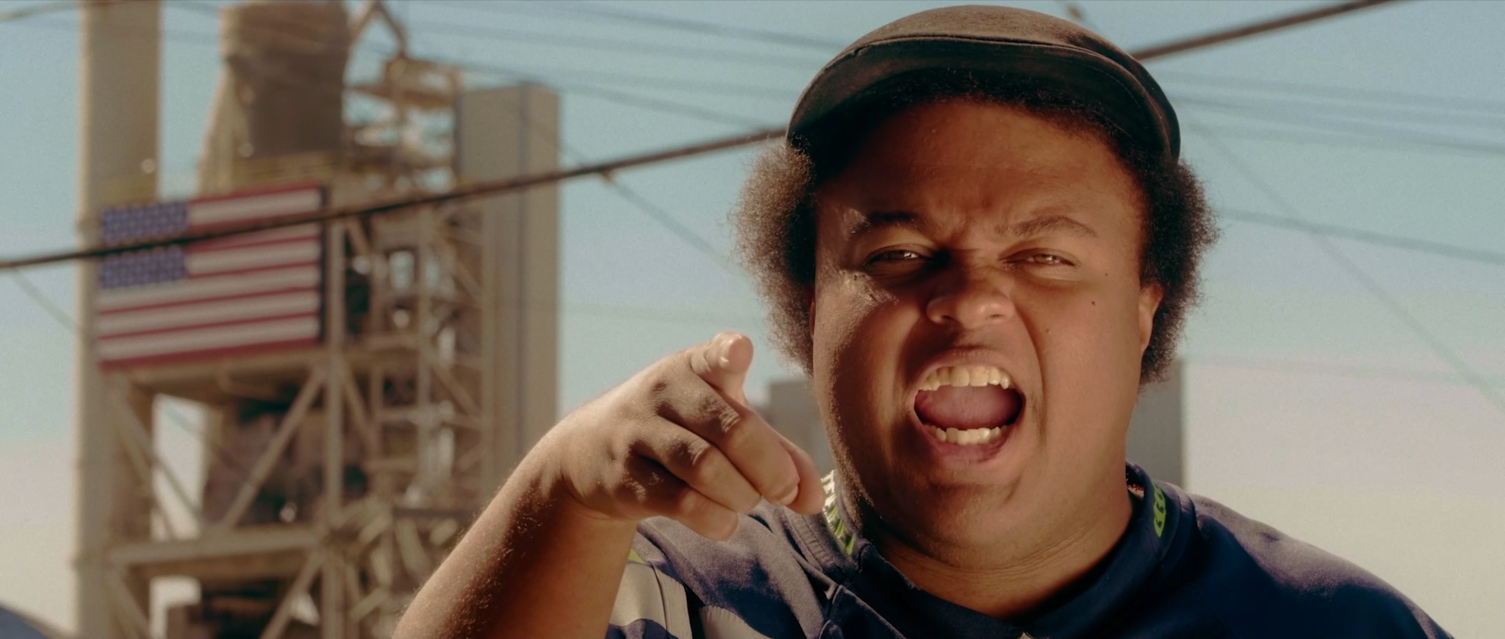 Close-up film still of a young person looking and pointing a finger directly at the camera; their mouth is open as if they are speaking with intensity. Power lines and a manufacturing plant with an American flag on its side stand in the background against a pale blue sky.