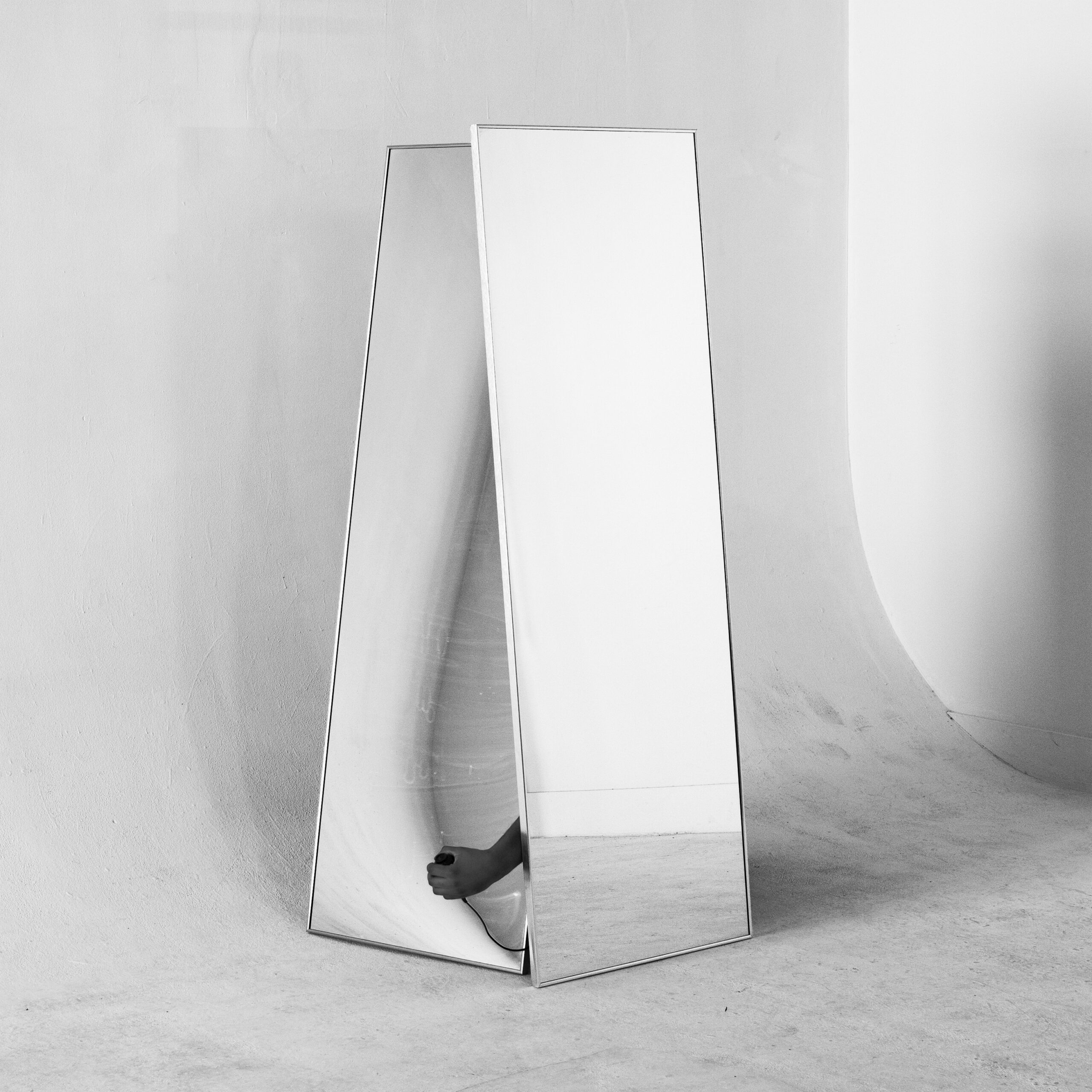 Black-and-white square photograph of a hand holding a shutter release cable, visible only through a gap between two tall rectangular mirrors leaning on each other.