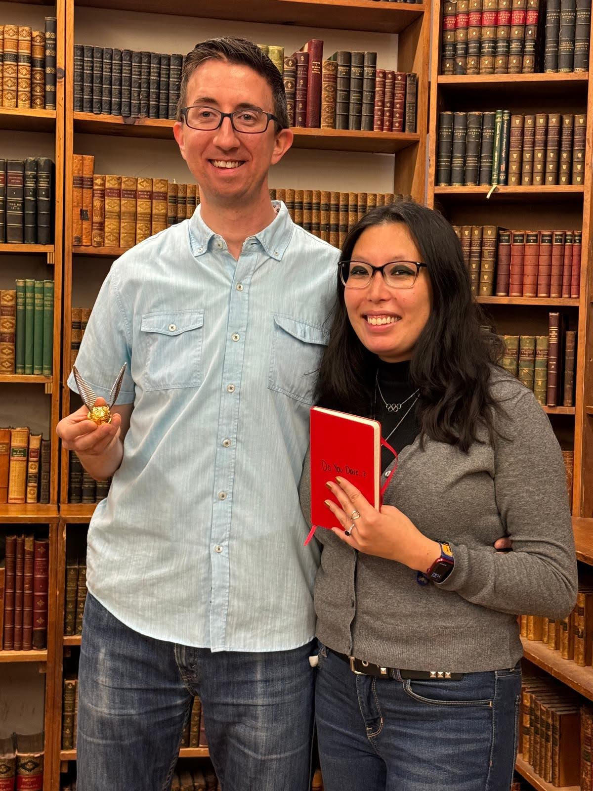 Two smiling people wearing glasses and blue jeans with their arms around each other, facing the camera in front of shelves with bound volumes, The person on the left holds a golden ball with silver wings; the person on the left holds a red notebook with the writing “Do You Dare?” across the front.