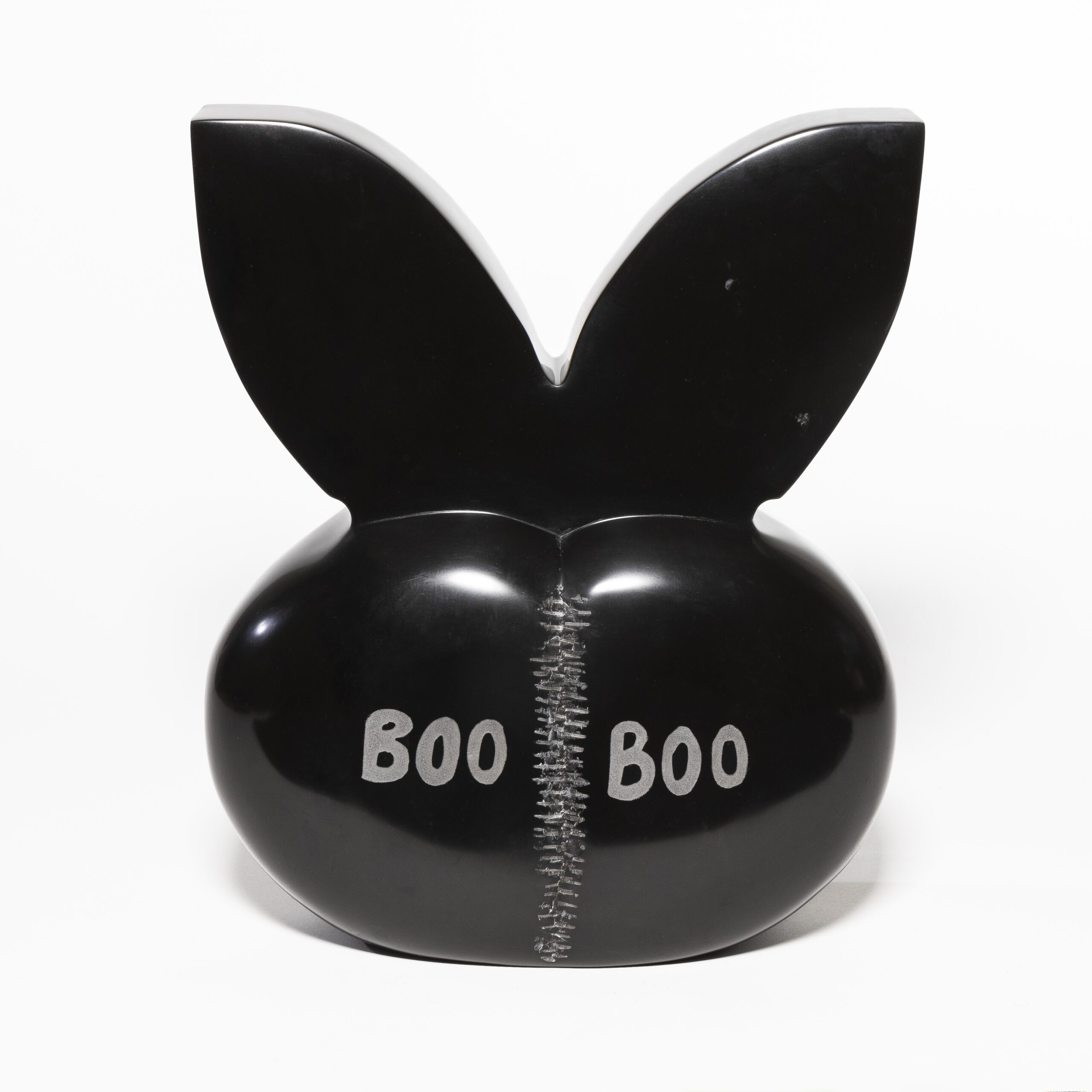 Set against a white backdrop, a black marble sculpture with rabbit ears and a silver stitch in its middle; on either side of the stitch, the text, “BOO”, printed side-by-side.