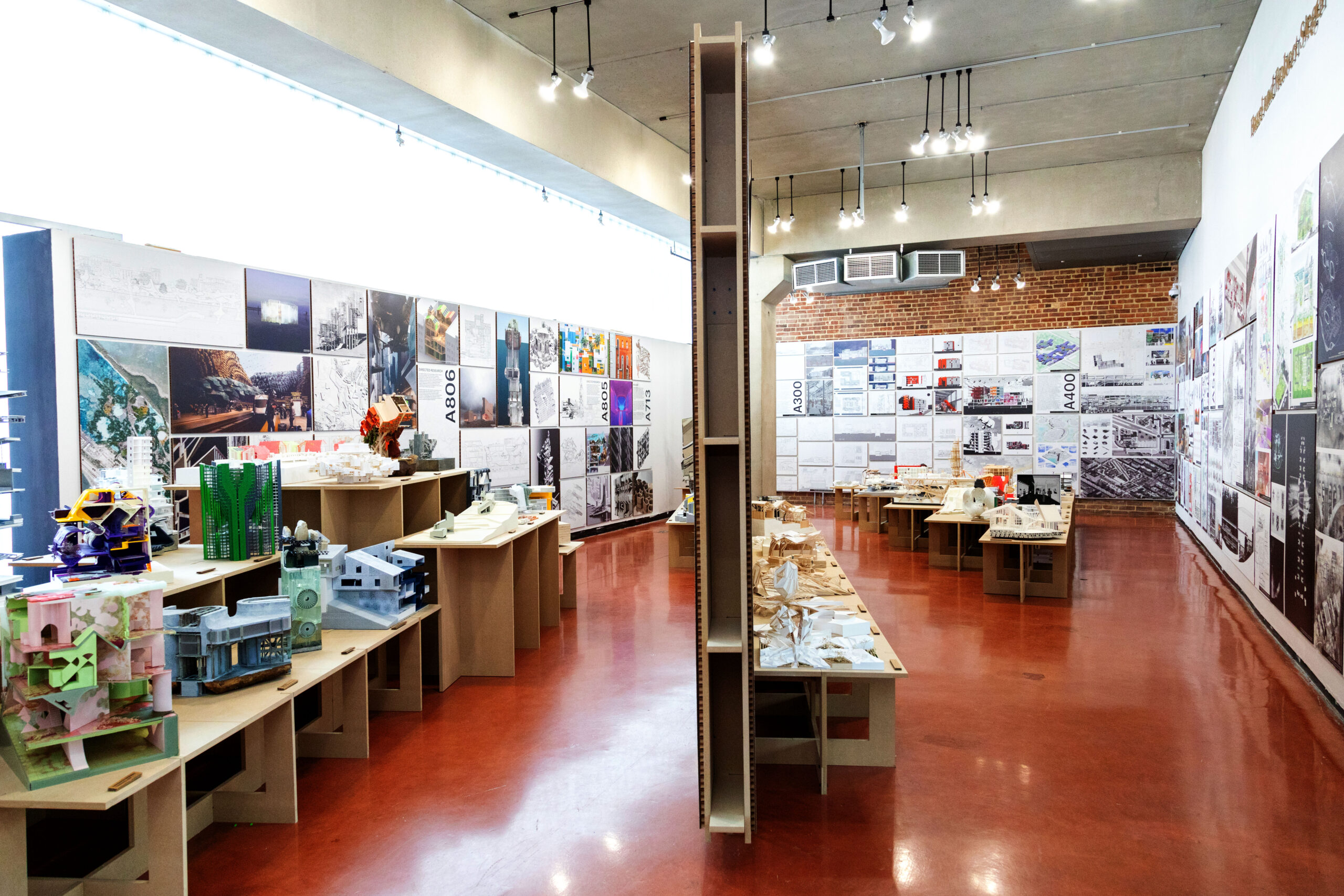 An exhibition space at Pratt Institute’s School of Architecture showcases dozens of architectural models and student projects. The room features high ceilings, red polished floors, and walls lined with detailed drawings, renderings, and photographs labeled by studio course numbers. Tables and shelves display intricate scale models made from various materials, reflecting the creativity and technical skill of undergraduate and graduate architecture students. Bright overhead lighting and natural light from large windows illuminate the space.