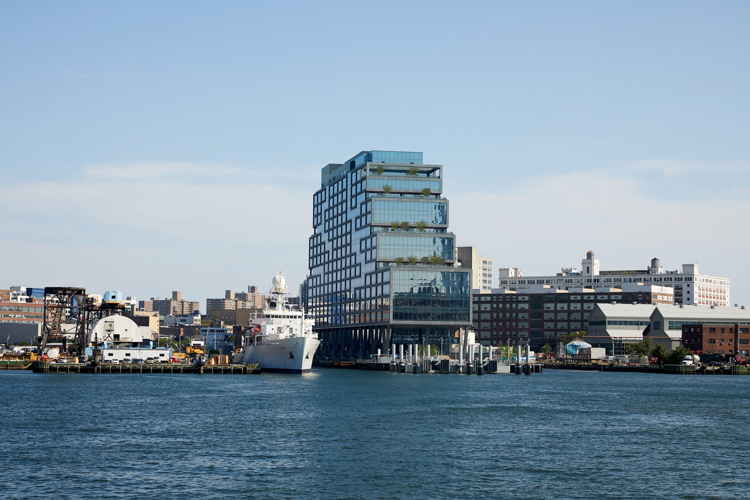 A modern, stepped glass building with reflective windows and terraces featuring greenery rises along the waterfront in an industrial urban setting. A large white ship is docked nearby, with warehouses and other industrial structures visible in the background. The sky is clear and blue, and the calm water in the foreground reflects the buildings and sky.