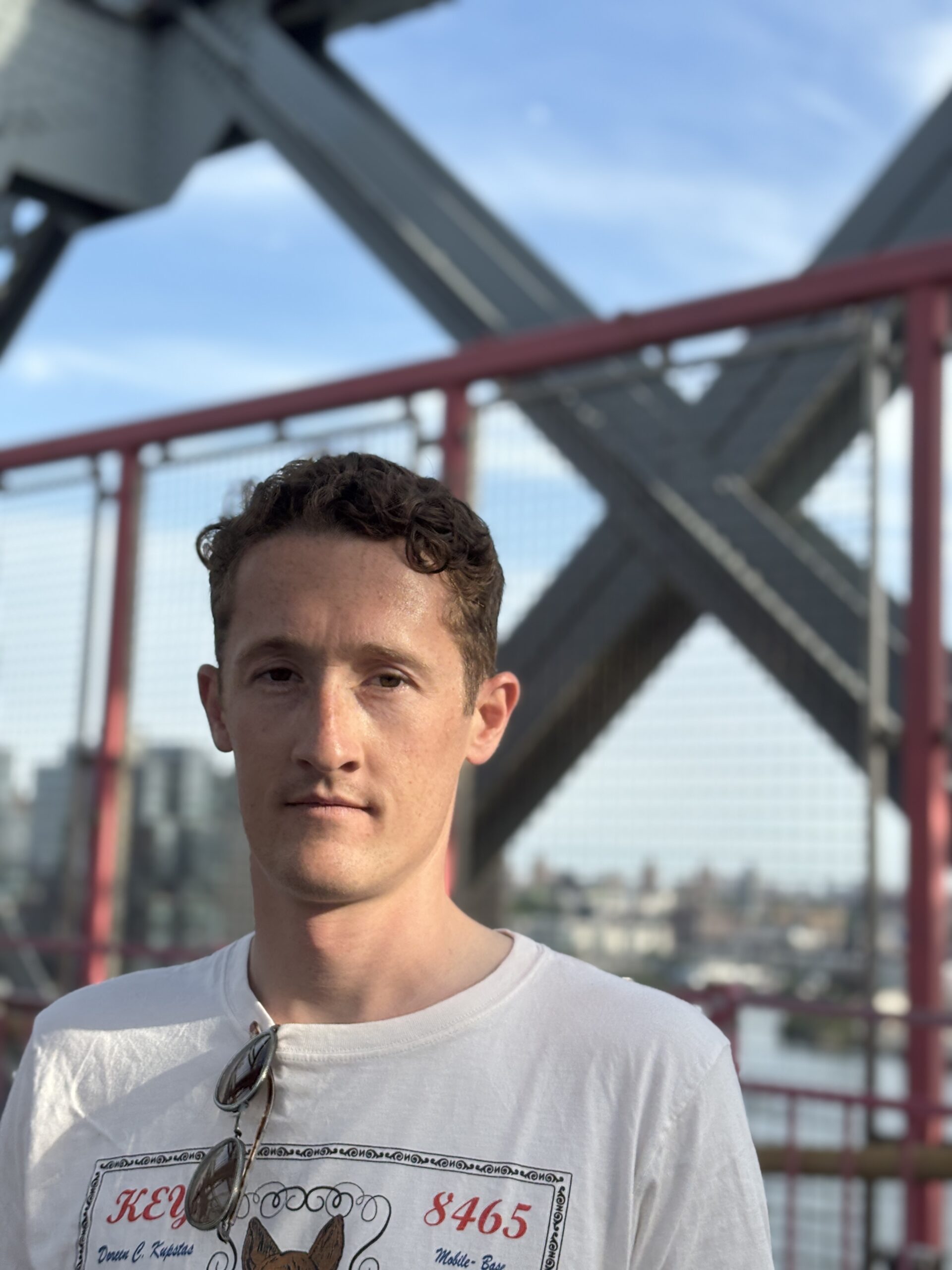A young man with short, curly brown hair and light skin stands on a bridge with a serious expression. He wears a white graphic t-shirt with red text and an illustration, and a pair of sunglasses hangs from the collar of his shirt. The background features a metal bridge structure with red railings and a cityscape visible in the distance under a clear blue sky. The soft lighting suggests it is late afternoon or early evening.