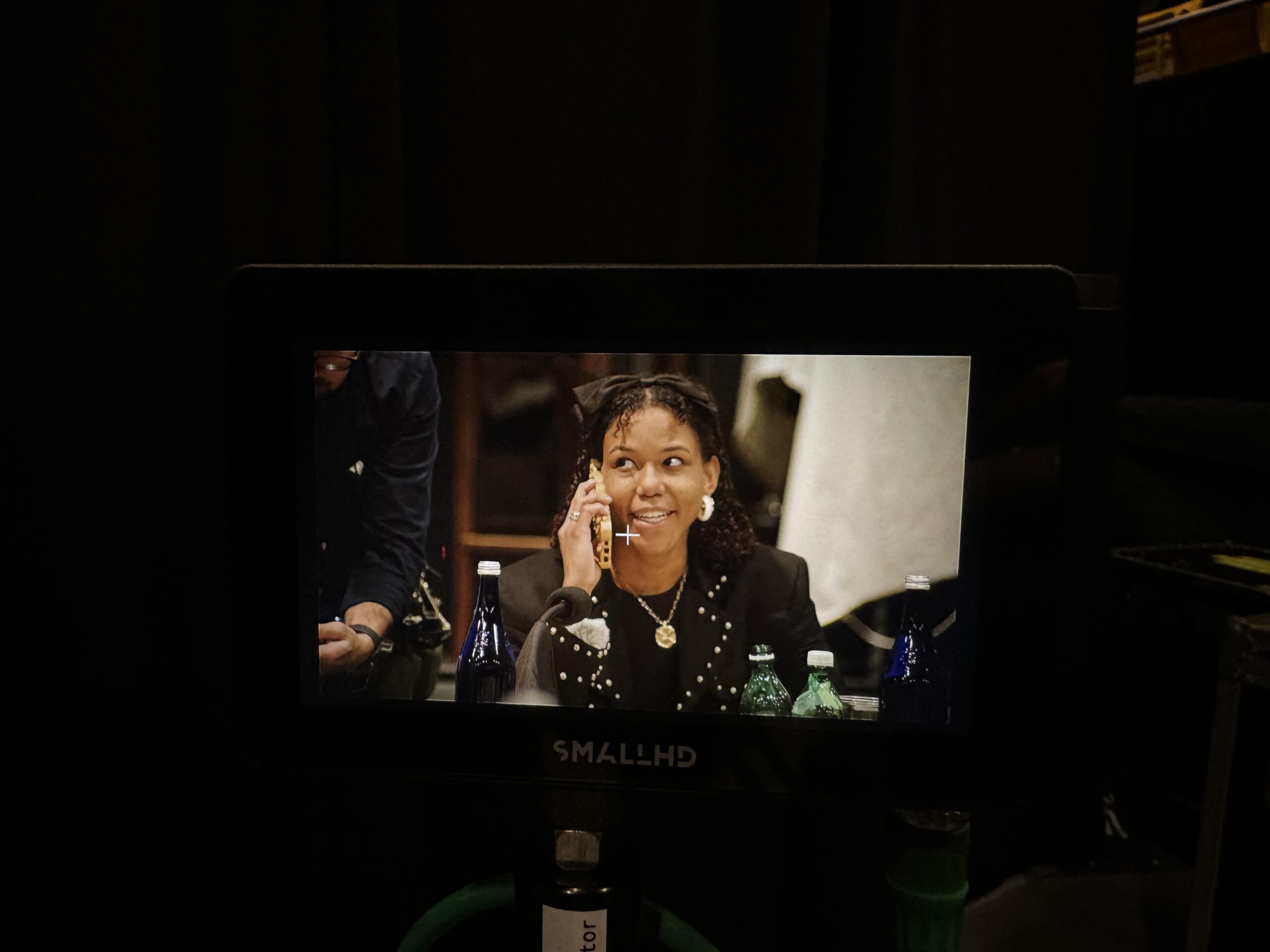 A woman is seen smiling while talking on the phone, captured through a monitor screen on a film or TV set, surrounded by bottles on the table and crew in the background.
