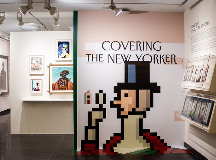 An exhibition space featuring a wall with the title "Covering The New Yorker" in bold lettering. Adjacent to the wall are various framed artworks, including portraits with colorful designs. One artwork features a character wearing a hat and holding a microphone, depicted in a pixelated style. The gallery has bright lighting and a clean, modern aesthetic.