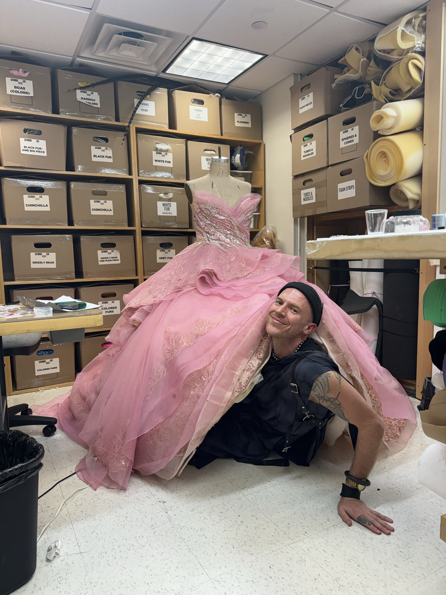A smiling man peeks out from underneath the voluminous layers of a vibrant pink ball gown on a dress form, in a storage room filled with labeled costume supply boxes.