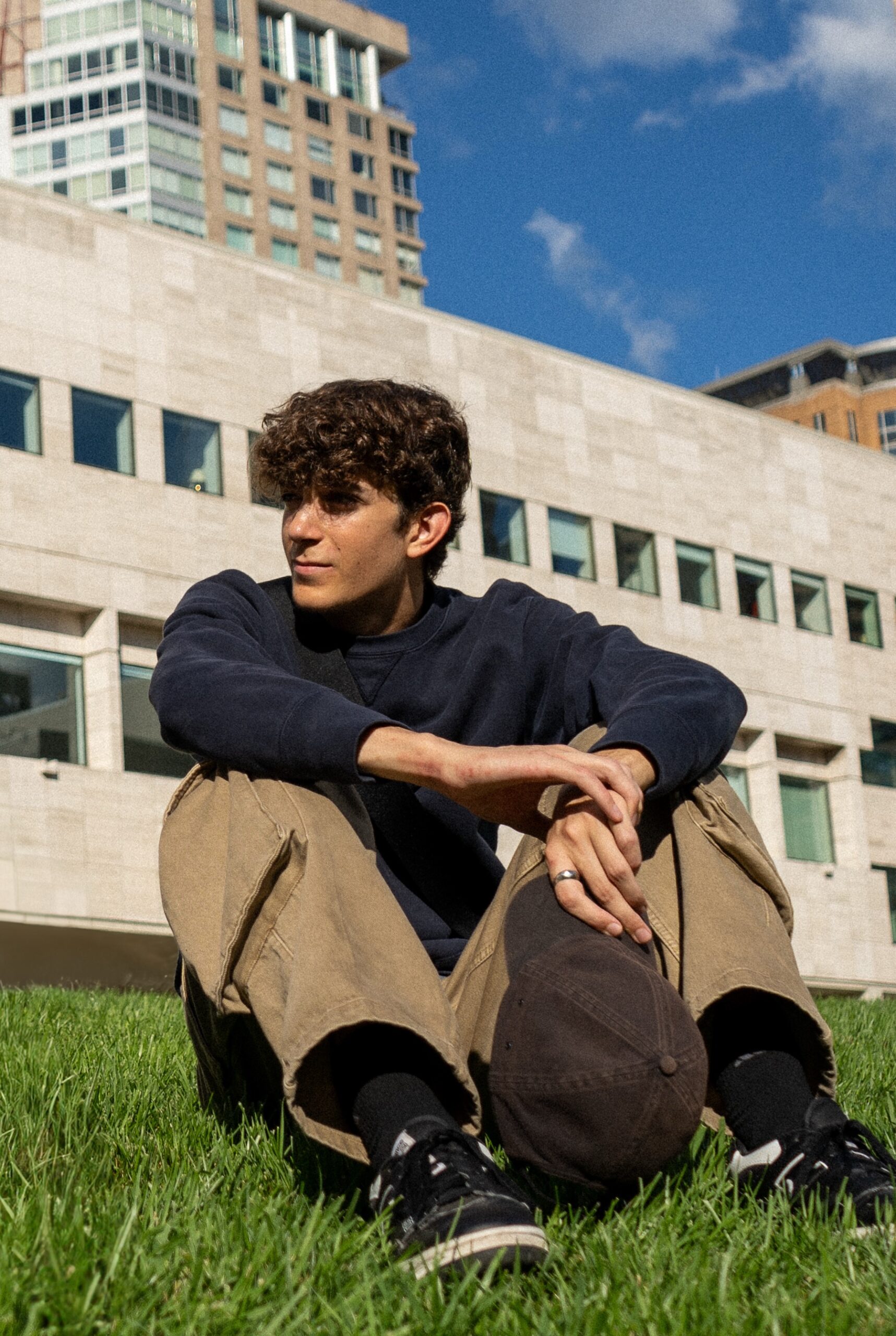 A young man sits cross-legged on a grassy lawn in front of a modern, light-colored stone building with rectangular windows. He is wearing a navy sweatshirt, khaki pants, black sneakers, and holds a dark baseball cap. Tall buildings rise in the background under a bright blue sky with scattered clouds.