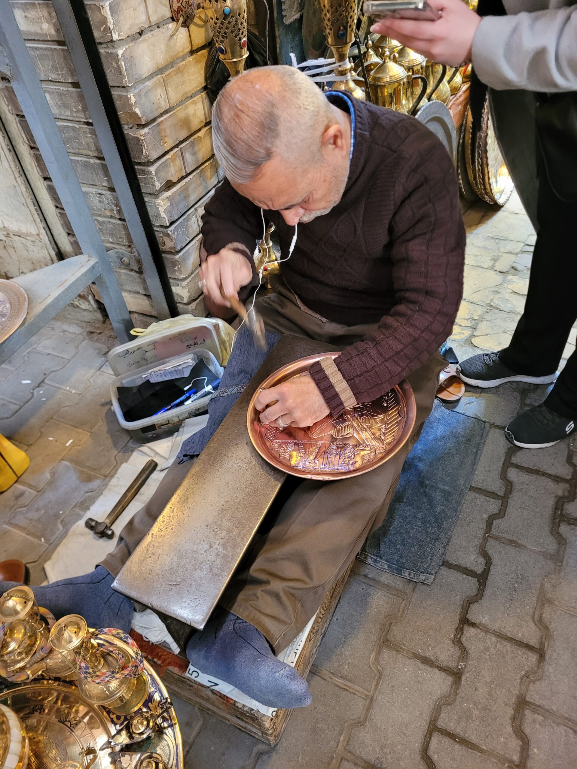 An elderly craftsman sits on a low bench working on a copper plate with a hammer and chisel. He is focused on engraving the surface while surrounded by various brassware in a traditional market setting.