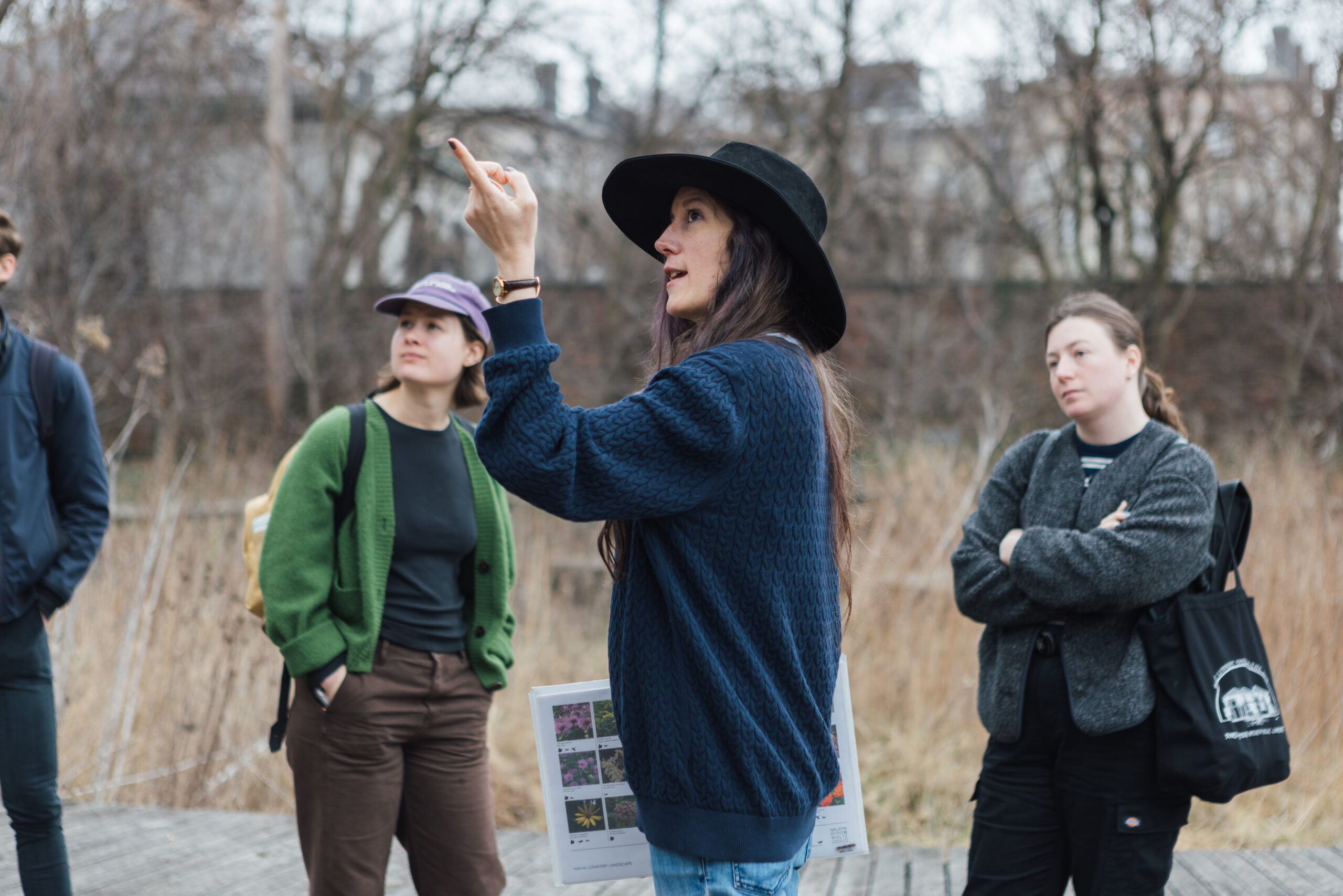 A group of students listens attentively during an outdoor tour at the Naval Cemetery Landscape. A woman in a black hat gestures while speaking, holding a plant reference guide, as others look on surrounded by tall grasses and bare trees in the background.