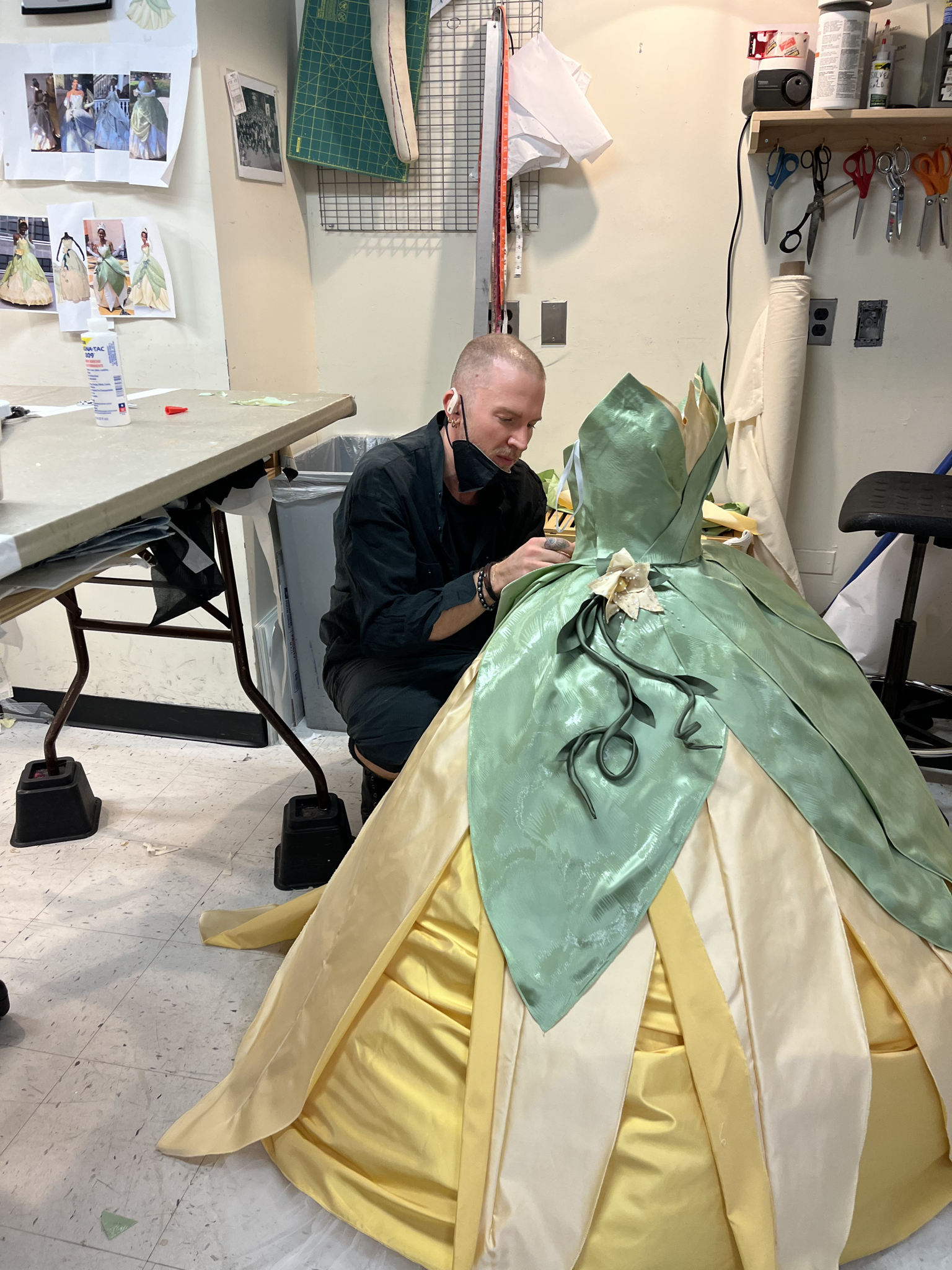 A costume designer, seated on a low stool in a studio, carefully works on a large, elegant gown inspired by Tiana from Disney’s The Princess and the Frog. The dress features layered yellow and pale green satin with petal-like panels and floral embellishments.