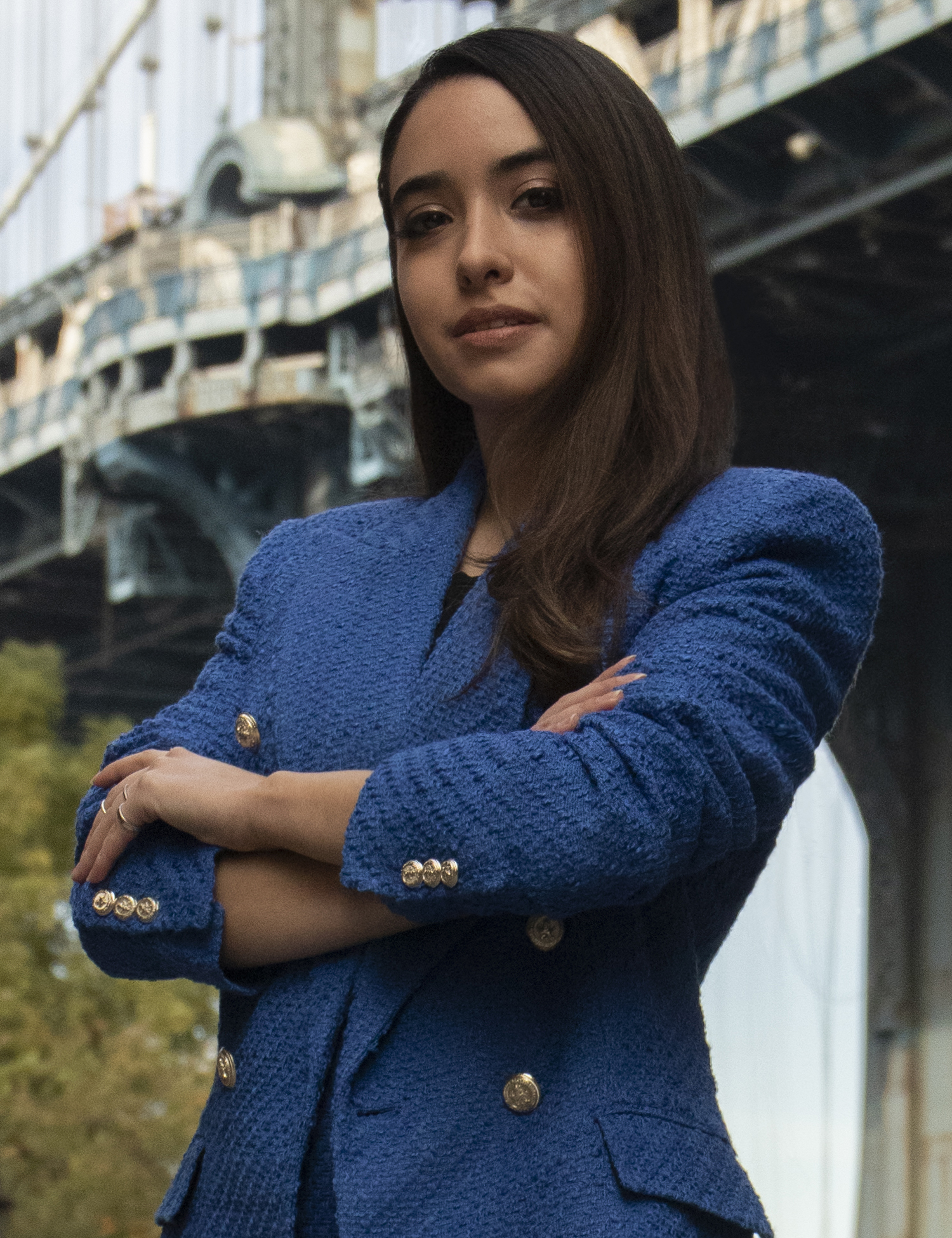 Headshot of Ordonez, who stands in a park with a view of a bridge in the background. She wears a blue blazer with gold buttons, arms crossed, looking at the camera with a neutral expression.