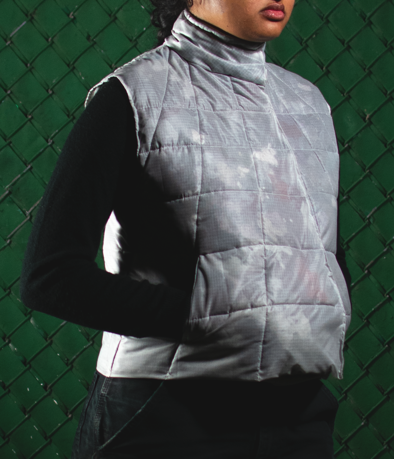 A person wearing a sleeveless, quilted grey vest made from translucent fabric filled with dryer lint. The vest has a high collar and angular seams. The background features a green chain-link fence.