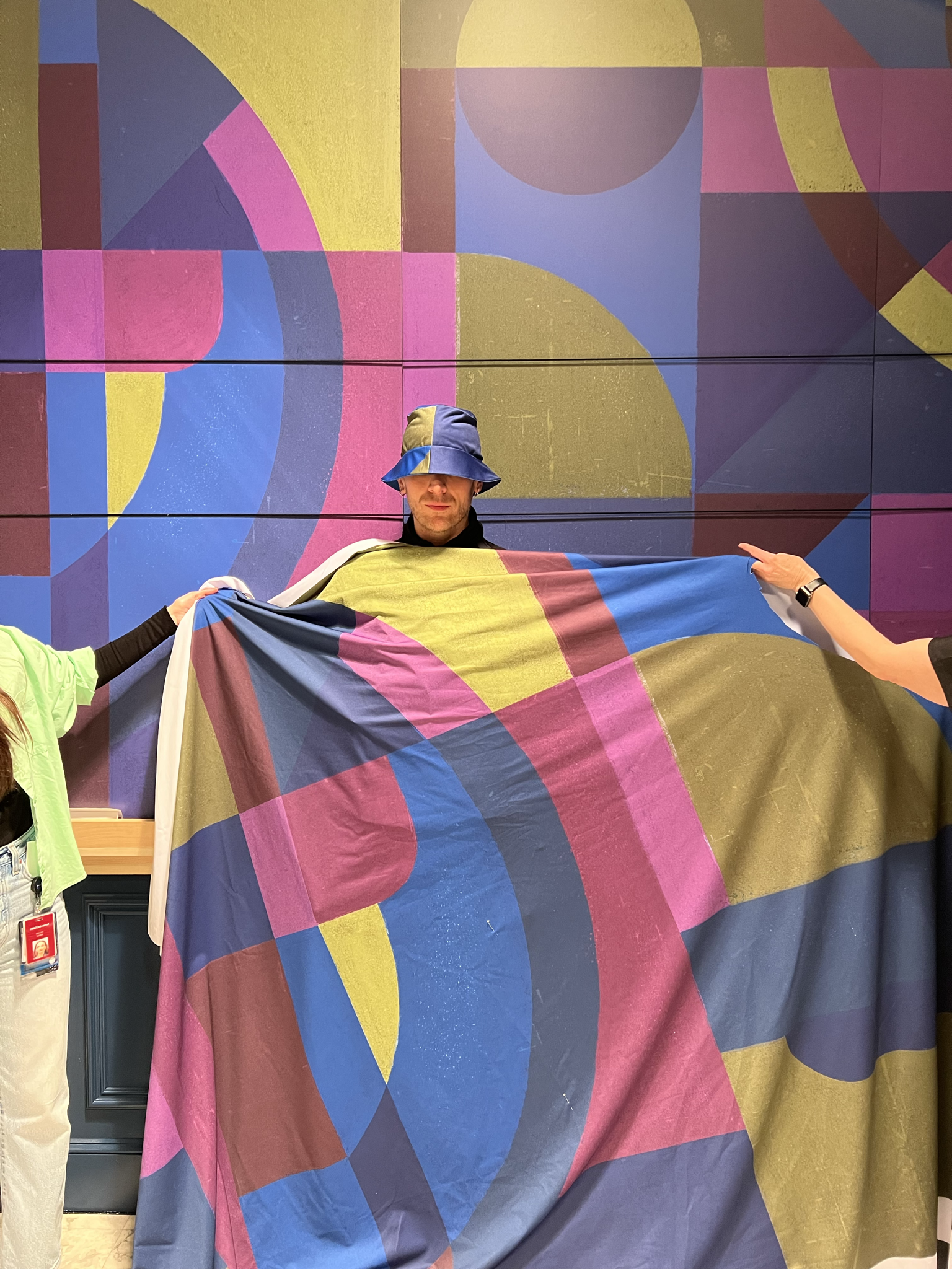 A man wearing a matching bucket hat and fabric wrap stands against a wall painted with a colorful, abstract geometric mural. The bold fabric draped around him echoes the mural's shapes and colors.