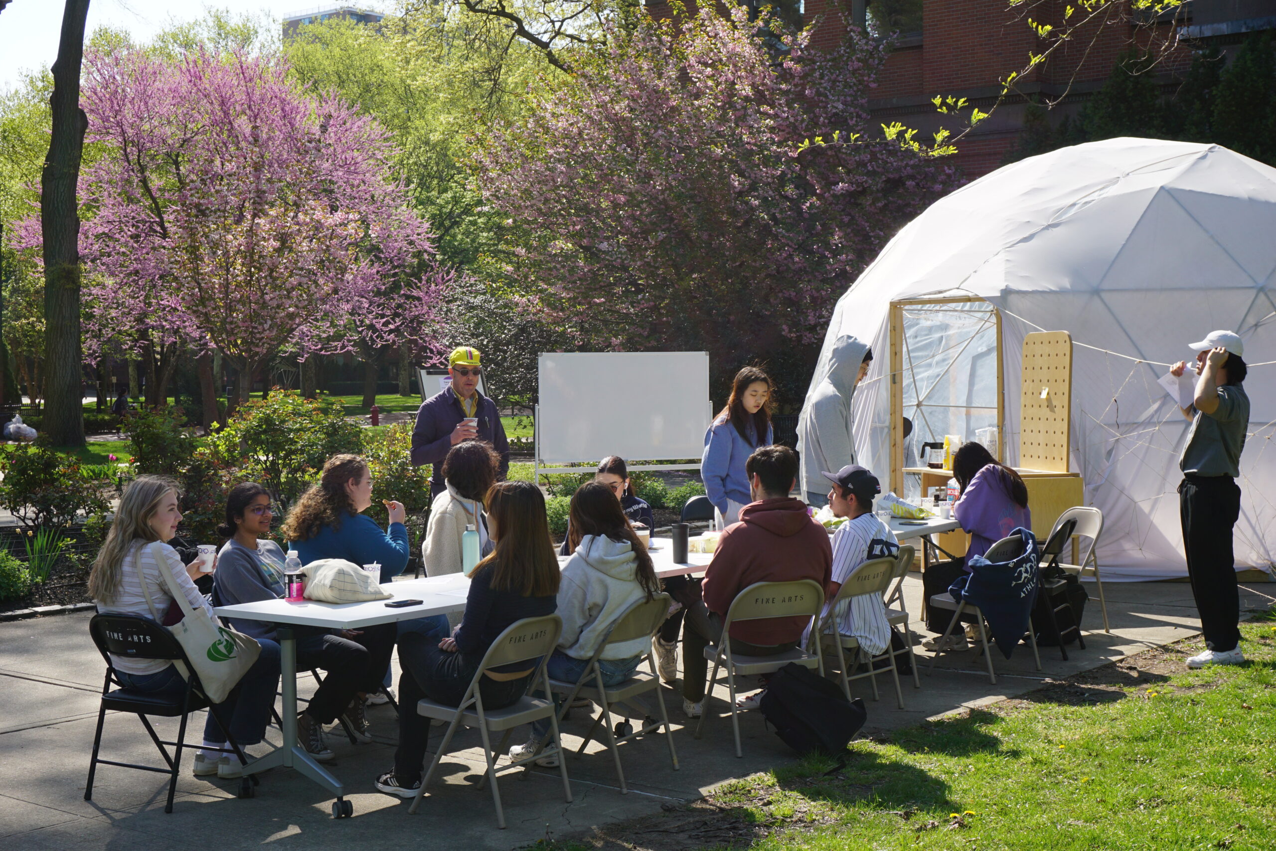 Students gather around a long table outside near a white geodesic dome while participating in a group discussion. The setting is lush with flowering trees and greenery, and one student appears to be giving a short presentation or speech to the group.