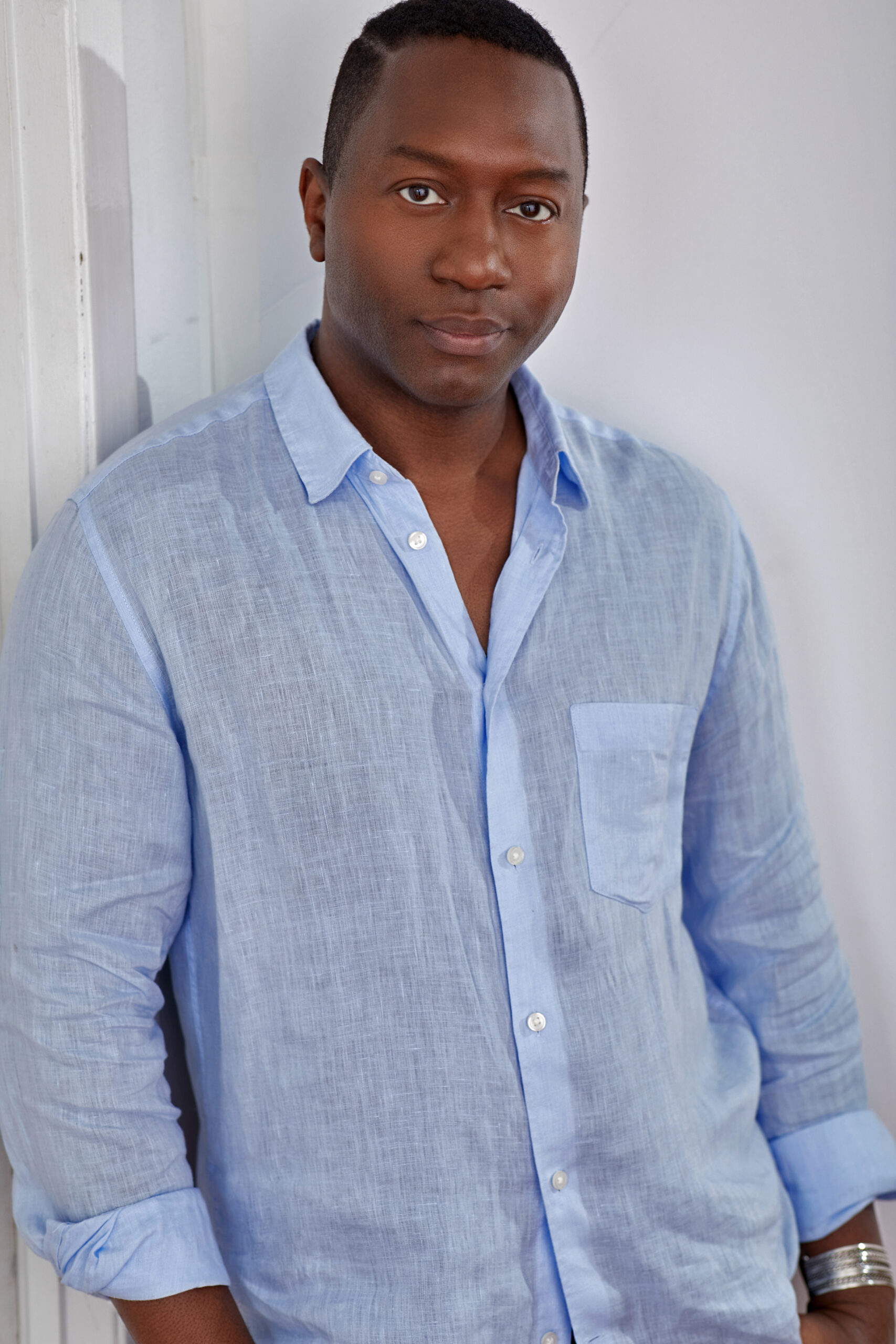 Thompson wears a light blue button-down and leans against a white wall, looking directly at the camera and smiling softly