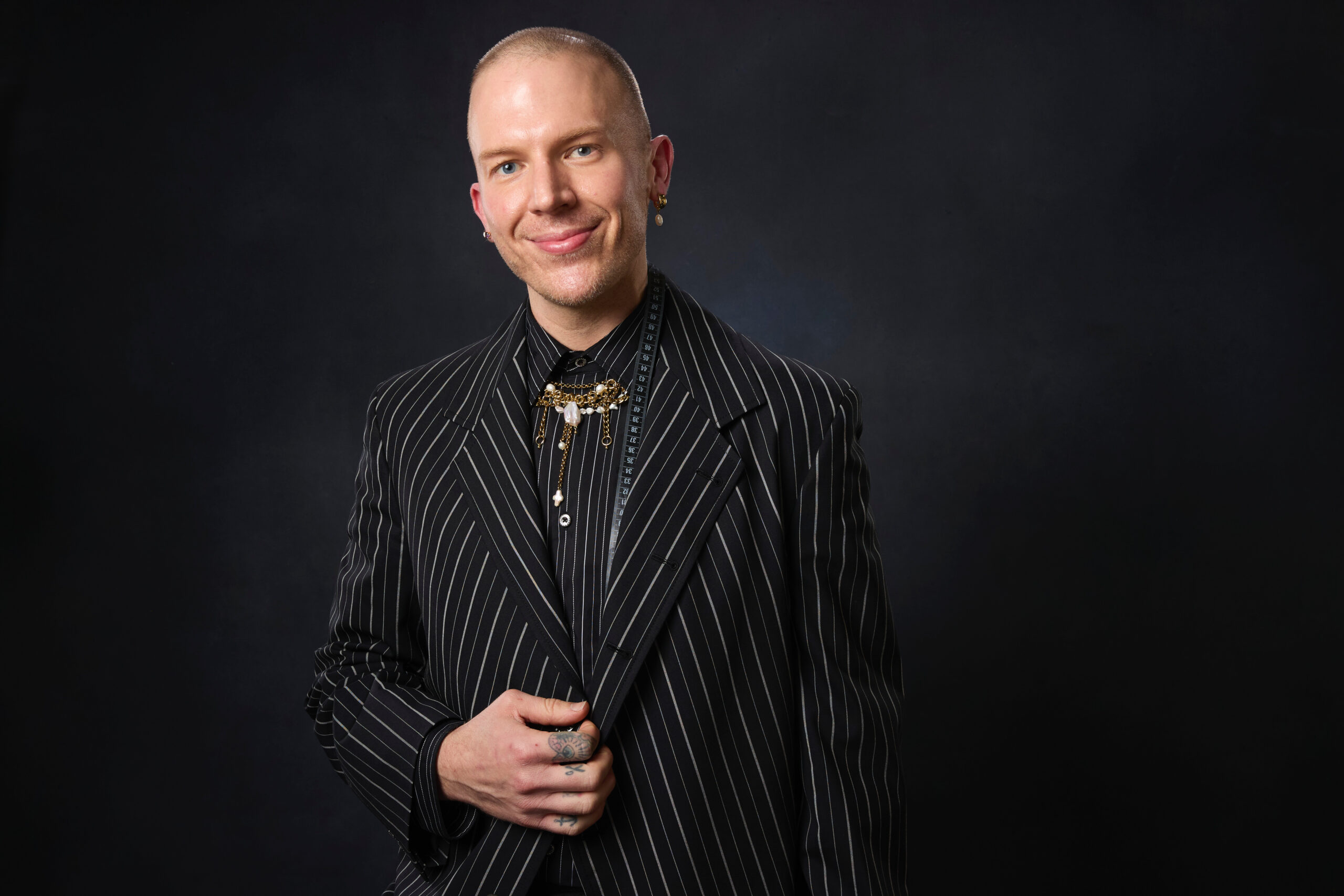 A professional portrait of a man wearing a pinstripe suit with a decorative brooch and earrings, standing against a dark background with a confident, friendly expression.