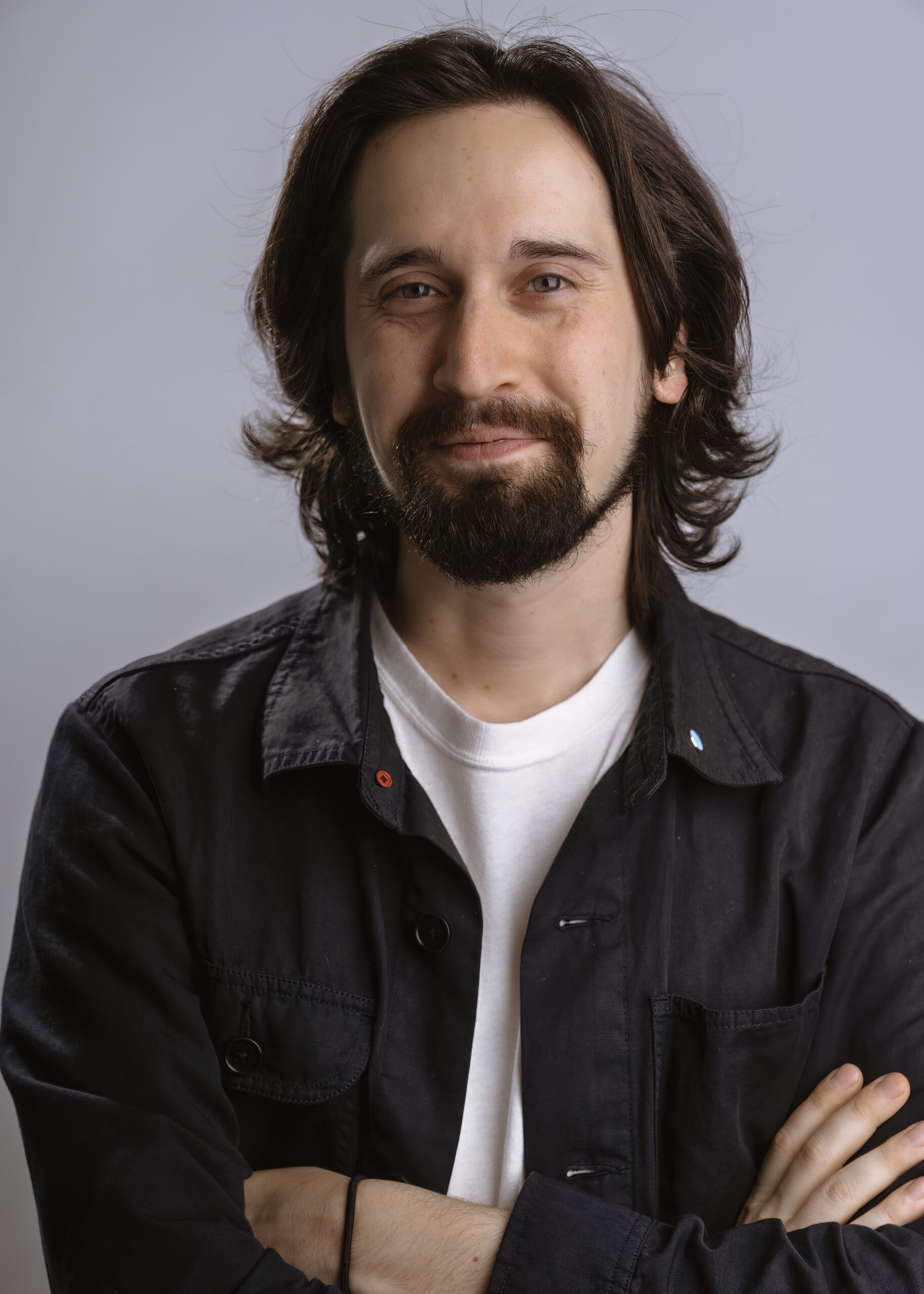 A headshot of Aaron Nesser against a light gray background. He is smiling at the camera with arms folded, wearing a white T-shirt under an open button-down.