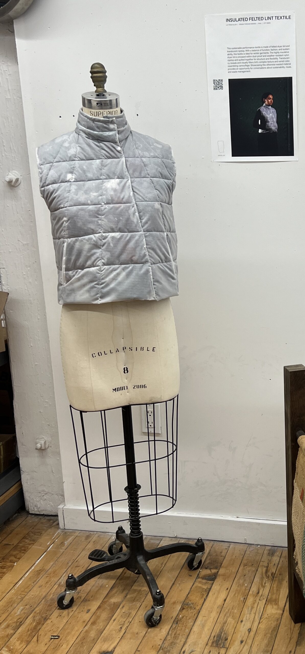 A padded vest displayed on a dress form in a studio. The vest is grey with a quilted pattern and is made from upcycled dryer lint encased in translucent fabric. A poster on the wall nearby provides information about the sustainable textile project.
