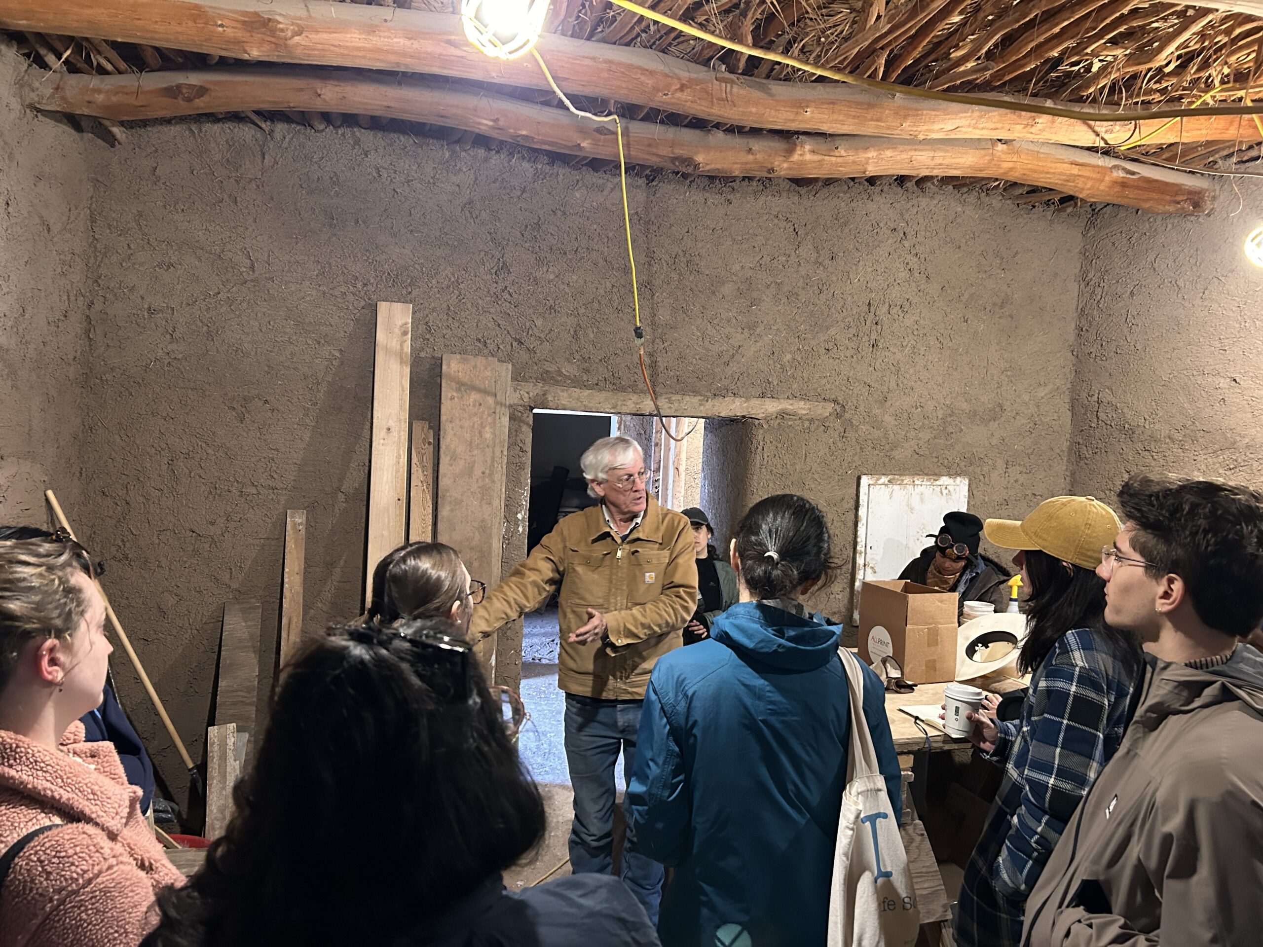 A group of people gathers inside a rustic adobe building with earthen walls and a wooden beam ceiling. An older man in a tan jacket stands at the center, speaking and gesturing with his hands while the group listens attentively. Lightbulbs hang from the ceiling, and wood planks lean against the walls, giving the space a workshop atmosphere.