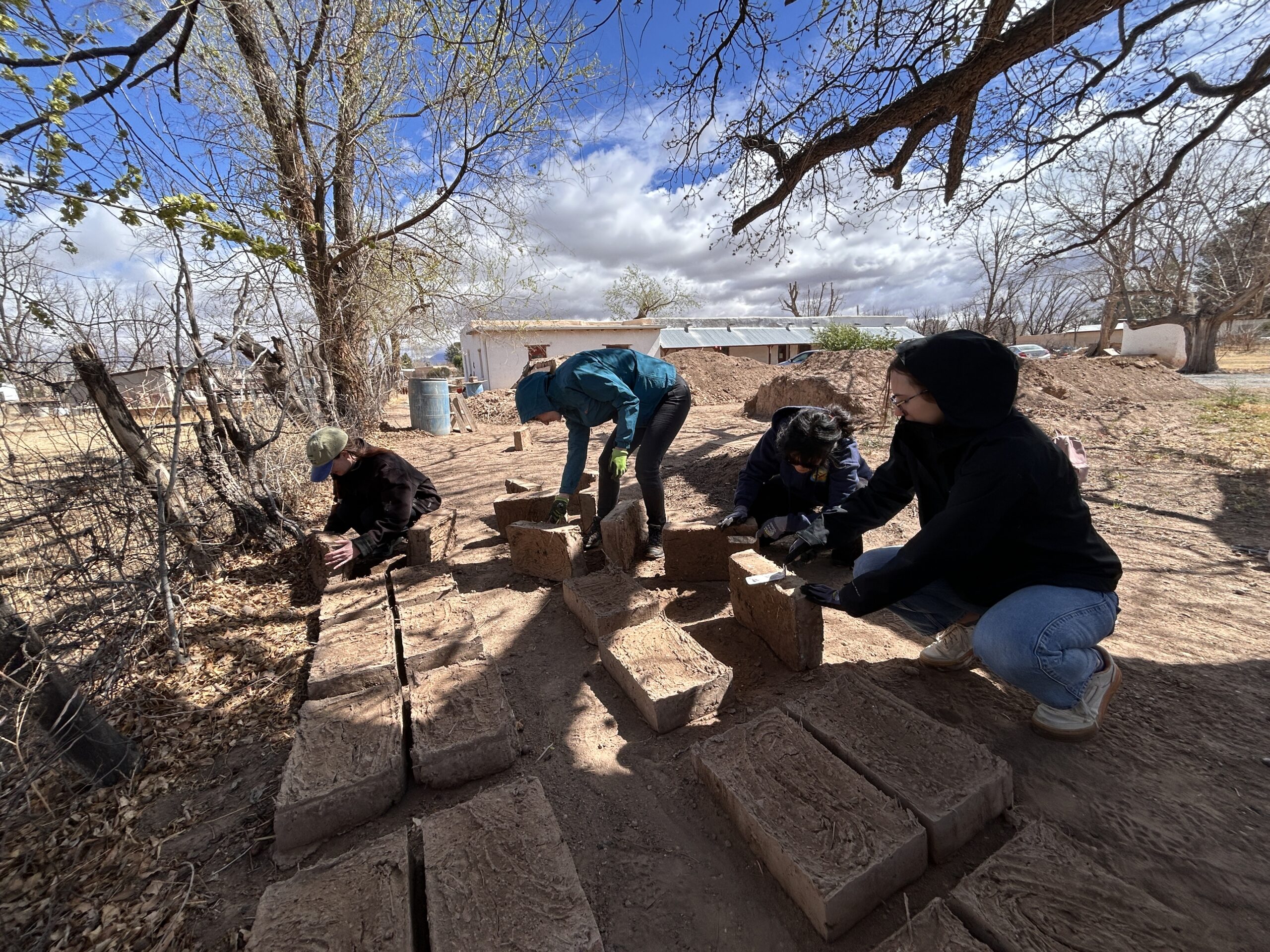 A group of people work outdoors under the shade of a tree, stacking and arranging adobe bricks on the ground. The scene is set in a rural area with dirt paths, scattered leaves, and a backdrop of bare trees, small buildings, and a bright blue sky with scattered clouds.