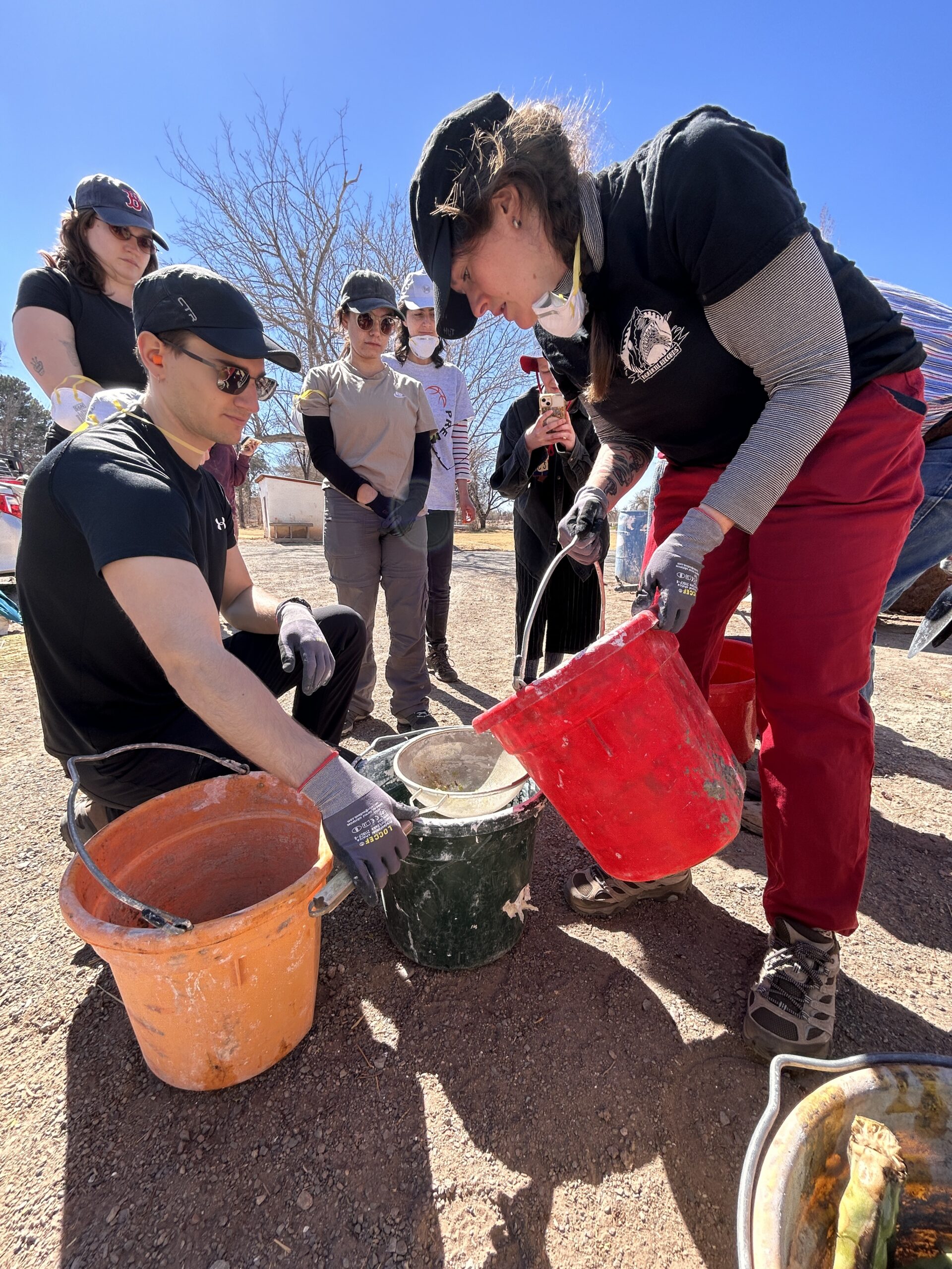 A group of people outdoors under a clear blue sky work with buckets and construction materials. A woman in a black hat, black shirt, and red pants pours a red bucket into a green bucket while a man wearing a black cap and sunglasses holds the bucket steady. Several others wearing gloves, masks, and casual work clothes observe the process.