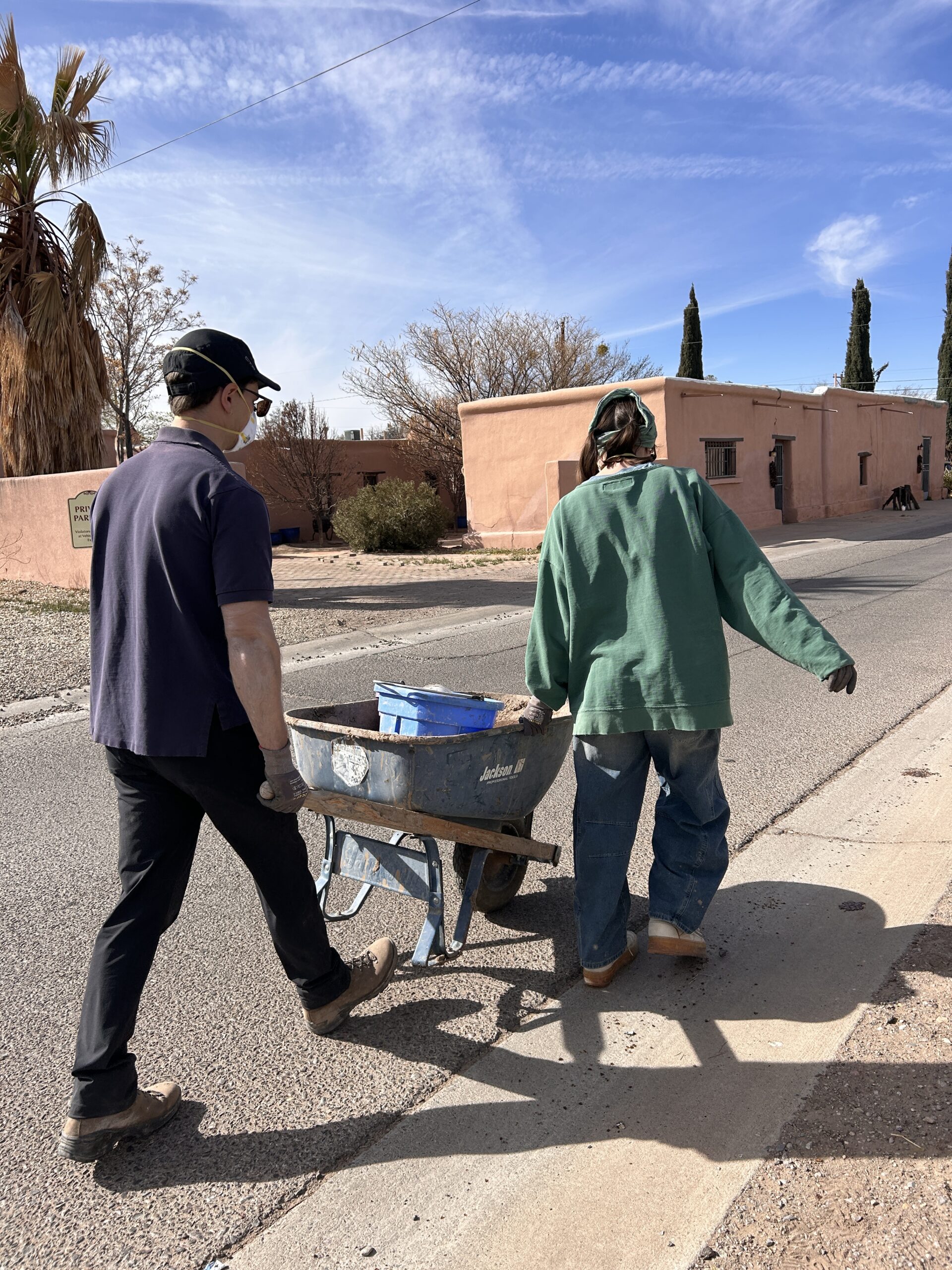 A person wearing a yellow baseball cap, safety glasses, mask, blue plaid jacket, pink pants, and tan boots uses a power drill to mix a substance in a bucket. They are working inside an earthen-walled building with two other people in the background also wearing masks and hats.