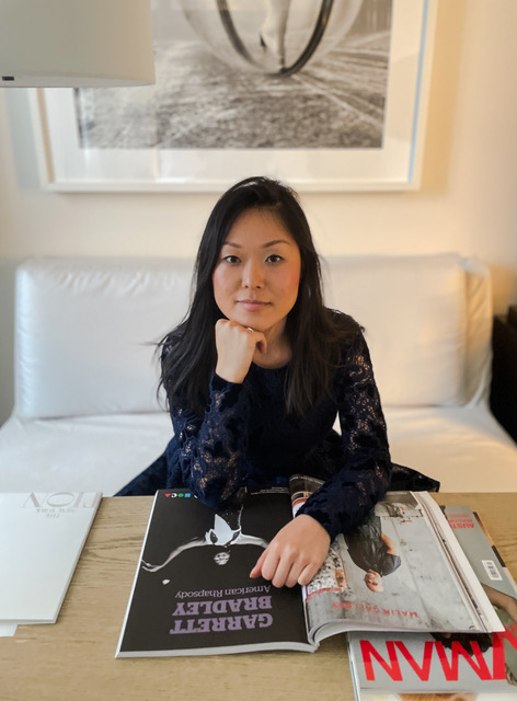 Liu, wearing a black top, sits in front of a desk with her head resting on one hand, her other hand laying over a magazine spread