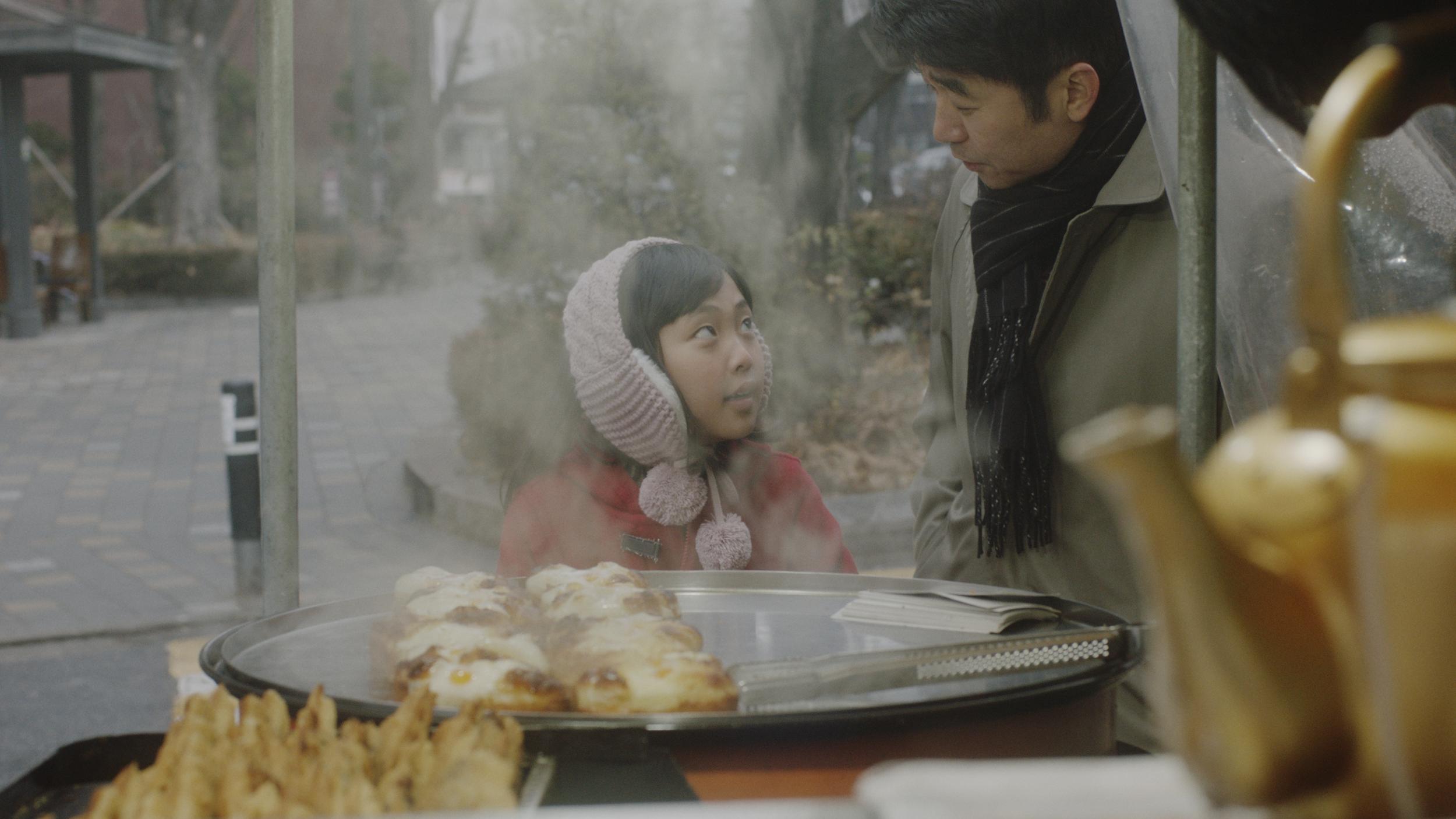 A young child and an adult speak to each other behind steaming plates of food in a grayish outdoor setting