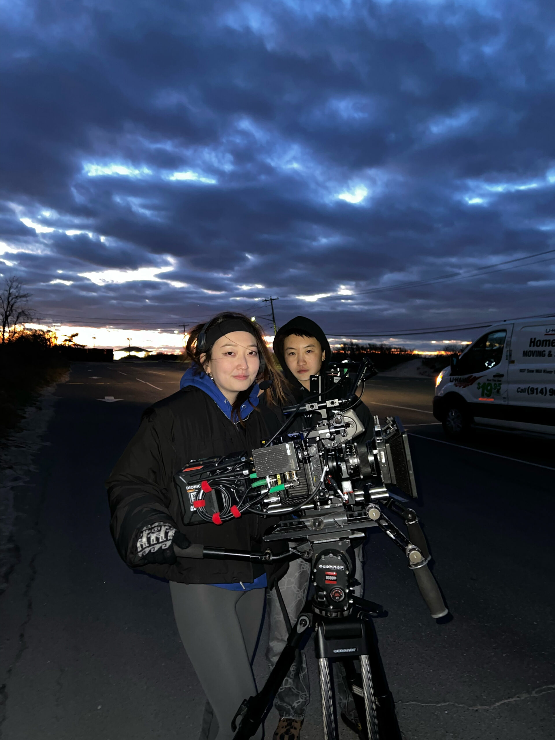 Eileen and Grace on set with video equipment alongside a road at dusk; the sky above them is cloudy and blue