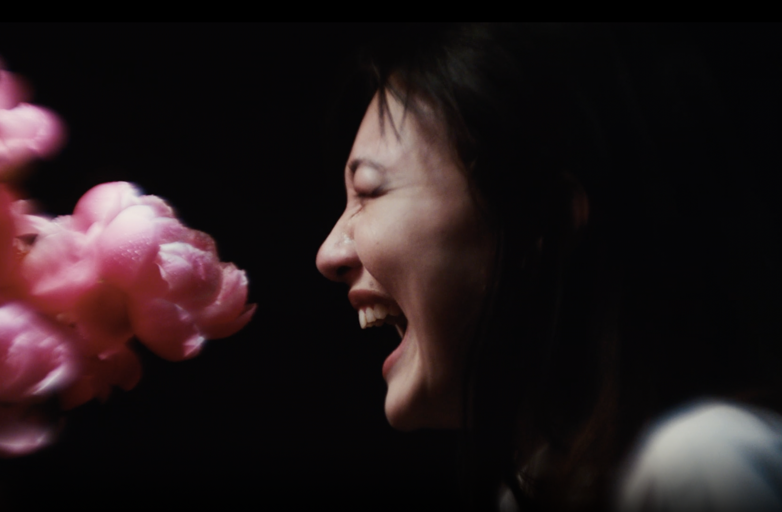 A person in center frame against a black backdrop laughs with their eyes shut and mouth open; across from them is a pink blooming flower in hazy focus