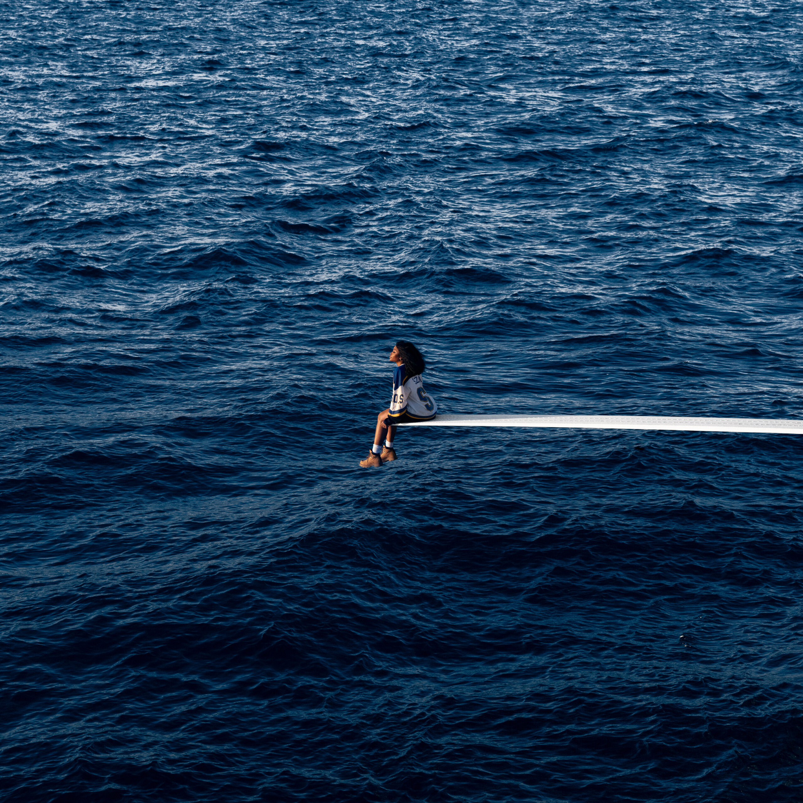 SZA, a woman wearing a jersey and Timberland boots, sits on the edge of a long diving board over a stretch of blue water, the sun shining on her face