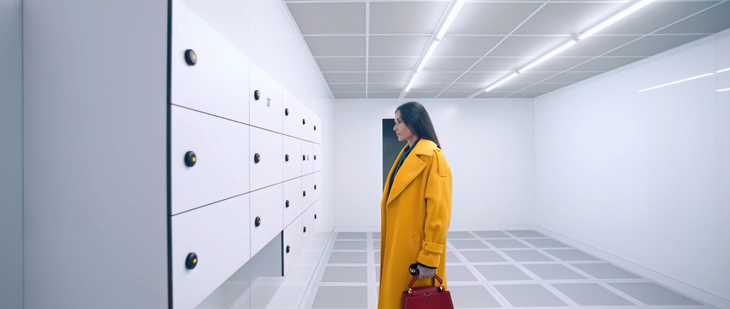 Demi Moore’s character wears a yellow coat, standing in a fluorescent-lit room with white walls and gazing intently at a wall of white lockers