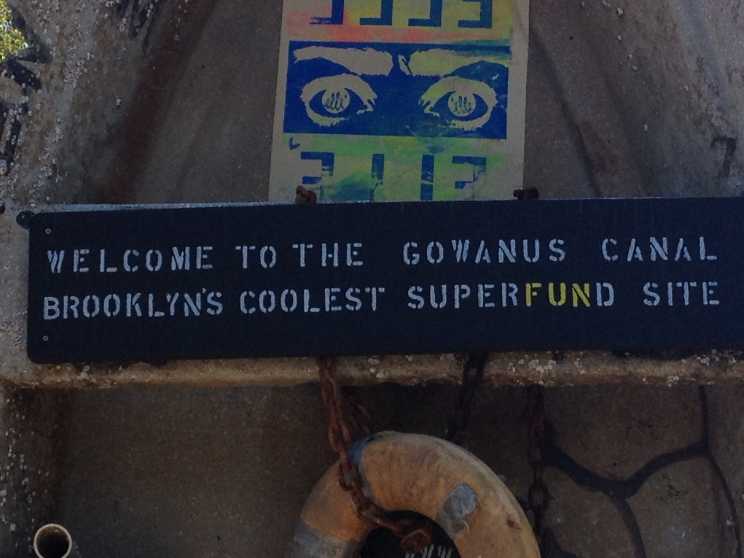 A close-up of a weathered black sign reading “Welcome to the Gowanus Canal: Brooklyn’s Coolest Superfund Site,” with the word