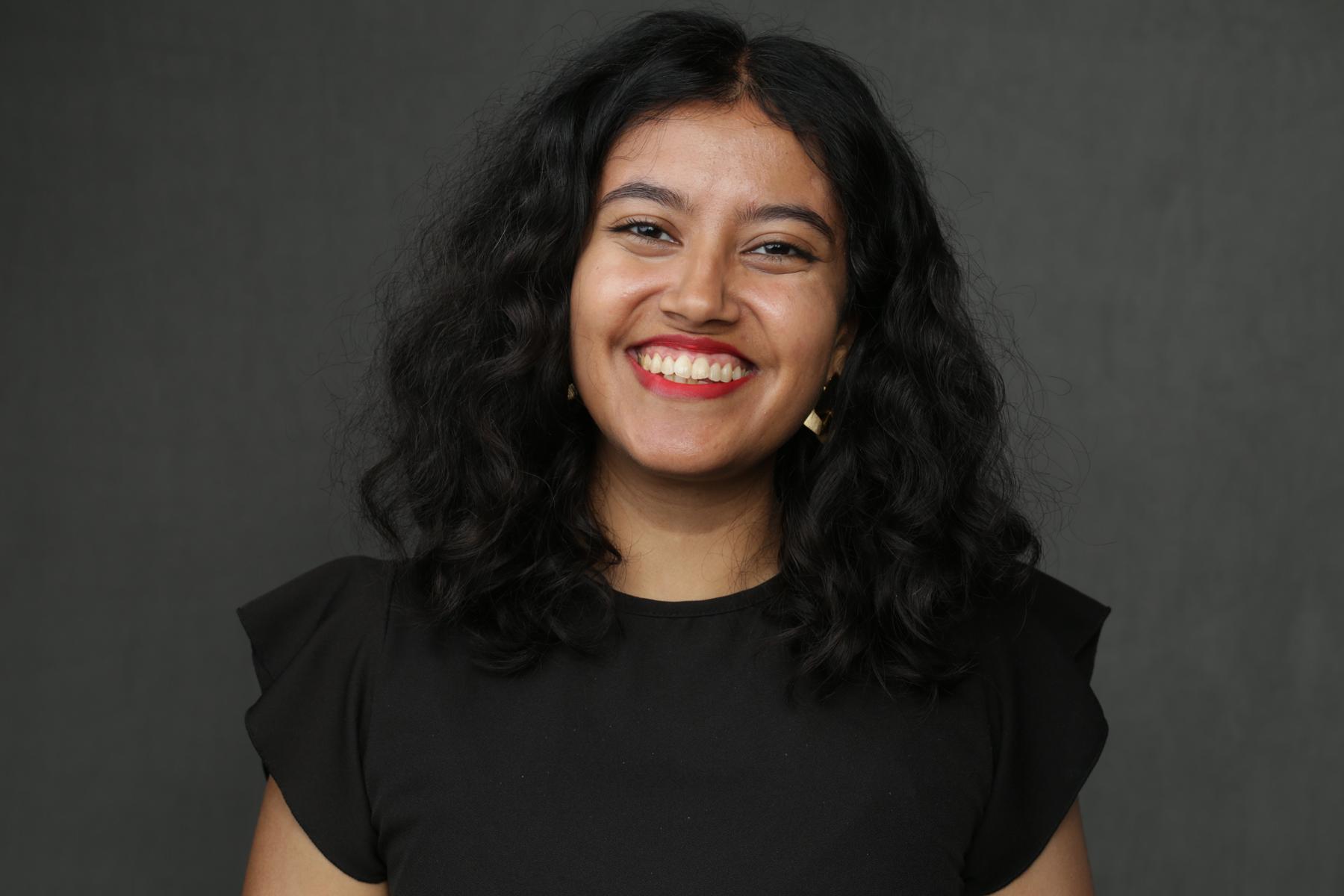 A headshot of Shikha Subramanian against a gray background. She wears a black shirt with ruffled short sleeves and smiles at the camera.
