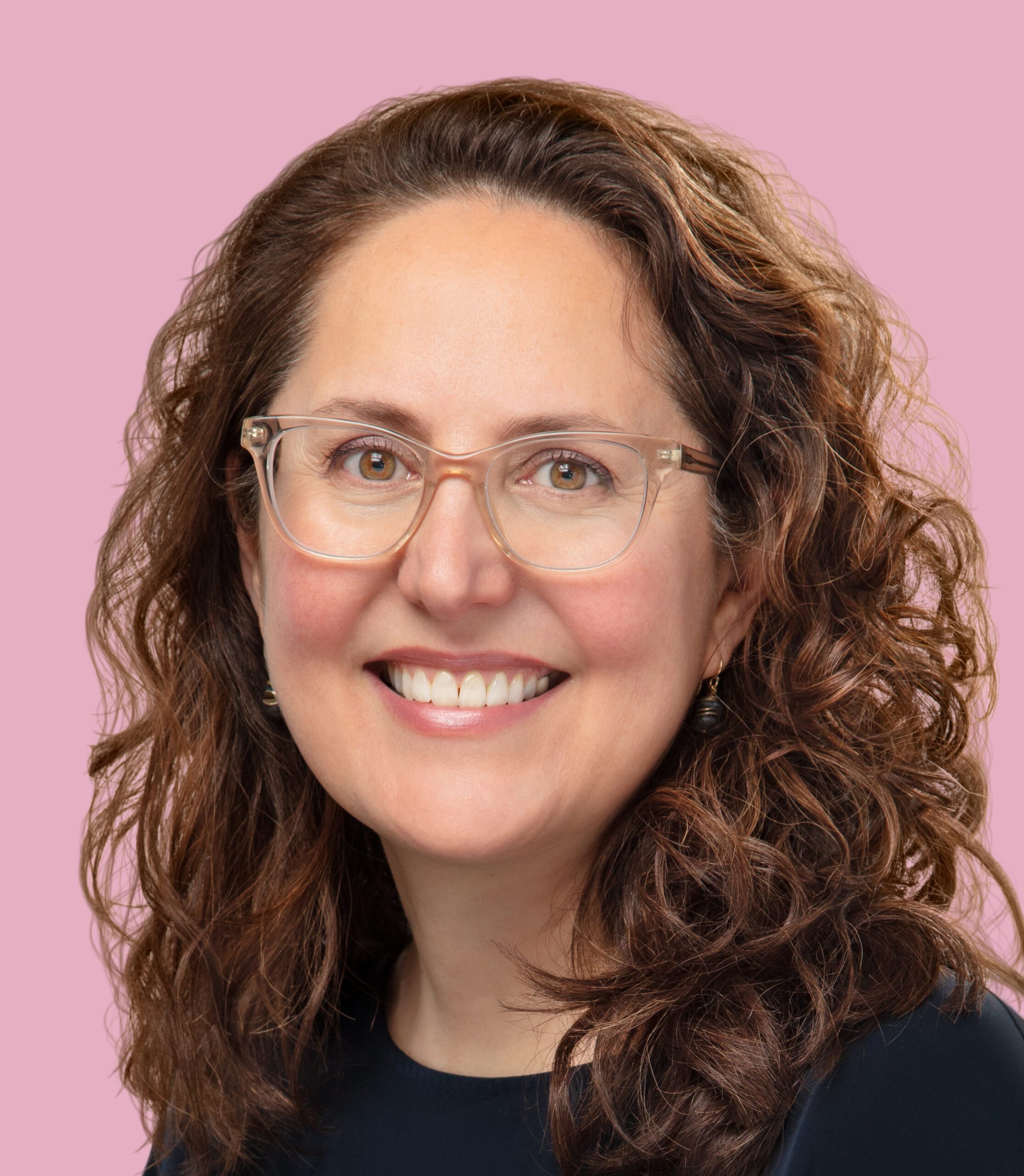 Close-up headshot of Dumery against a light pink backdrop. She wears clear-framed glasses and a black shirt, smiling at the camera.
