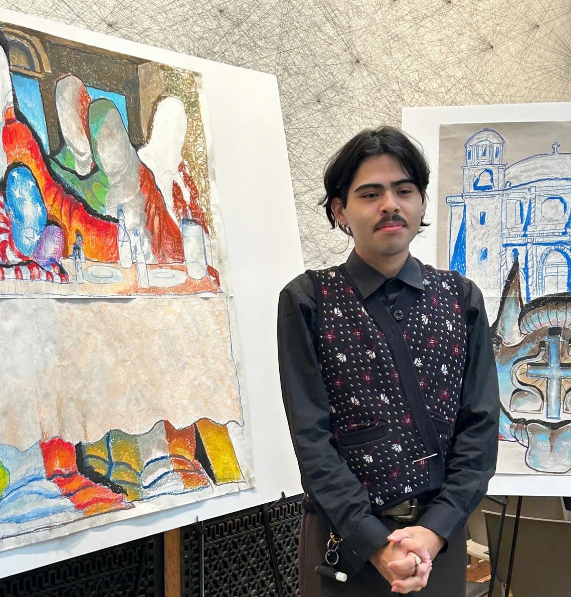 Angel Ortega stands beside two large, colorful drawings — one of the Last Supper and another of a historic cathedral — displayed on easels in a gallery setting.