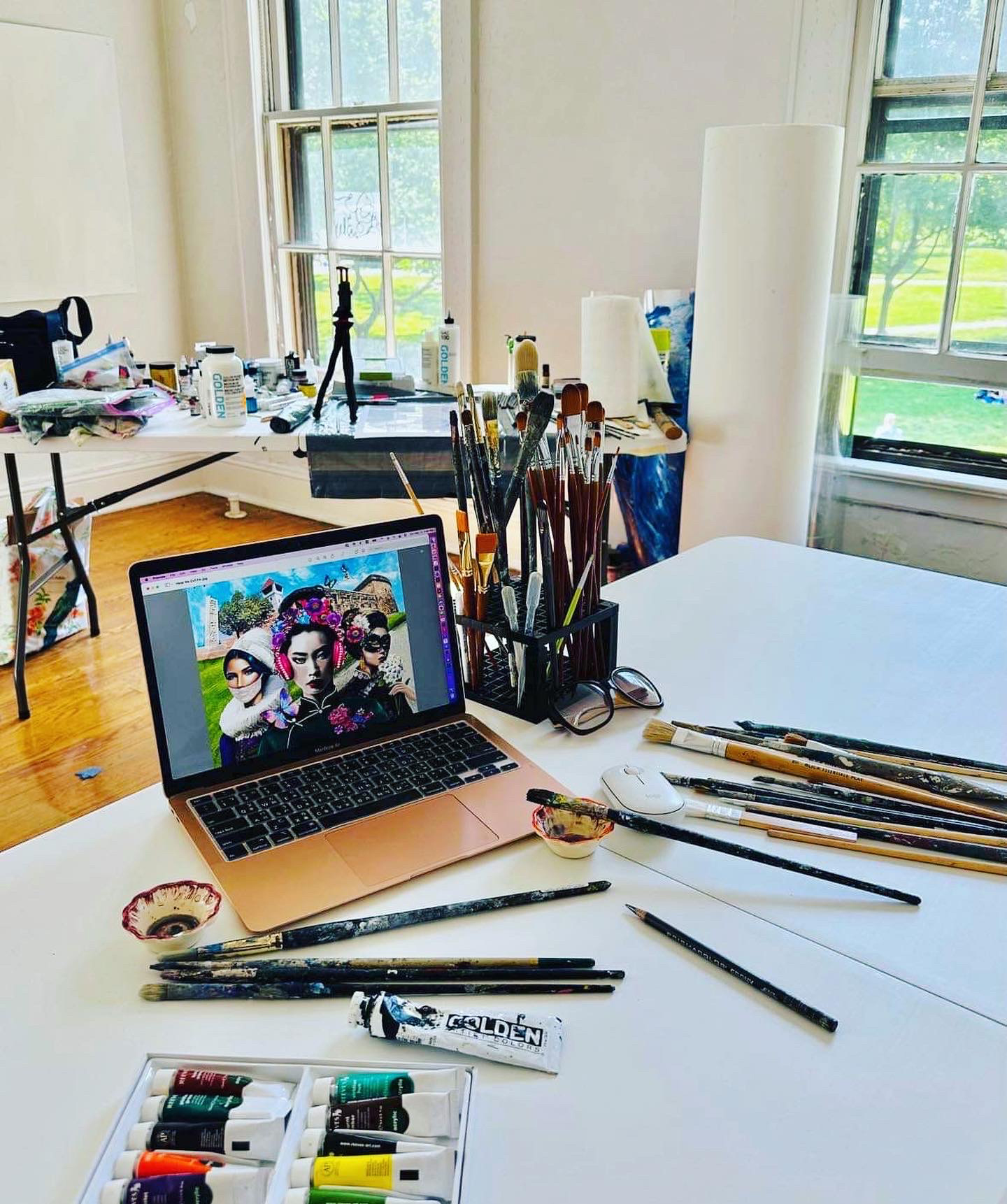 A creative workspace with tables covered in art supplies, brushes, and a laptop displaying digital artwork of three women. The setting is a well-lit room with wooden floors and large windows overlooking a green park, suggesting an artist residency or studio environment.