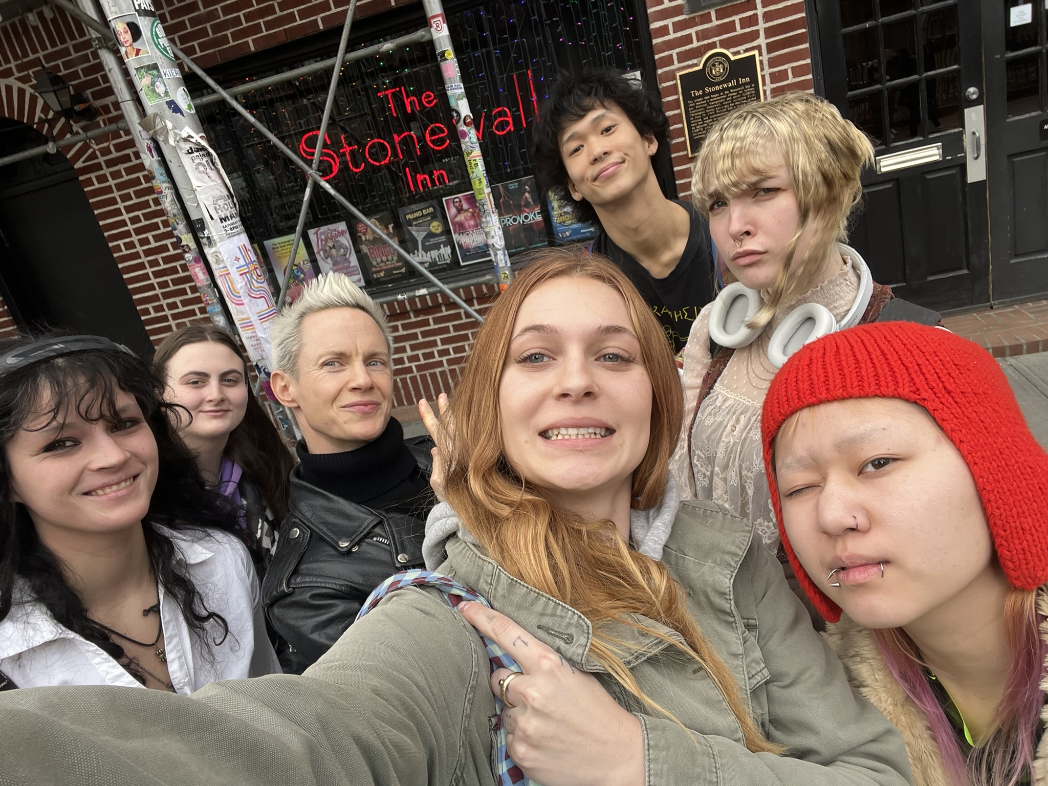 A group of seven people smiling and posing for a selfie in front of The Stonewall Inn, a historic LGBTQ+ landmark in New York City. The background shows the red brick exterior, neon sign, and posters on the wall.